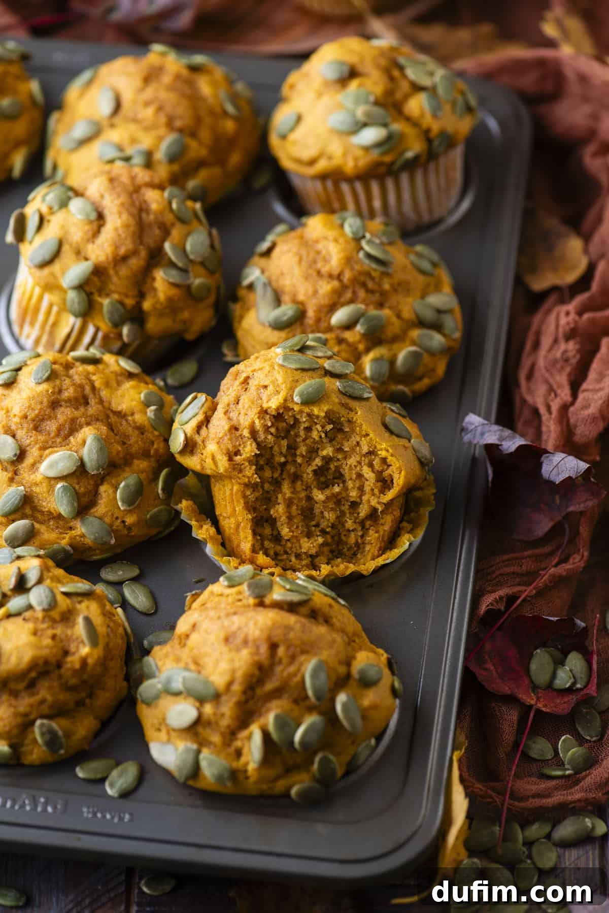 pumpkin muffins topped with pumpkin seeds in a muffin pan sitting on a dark orange towel, surrounded by pumpkin seeds and fall leaves