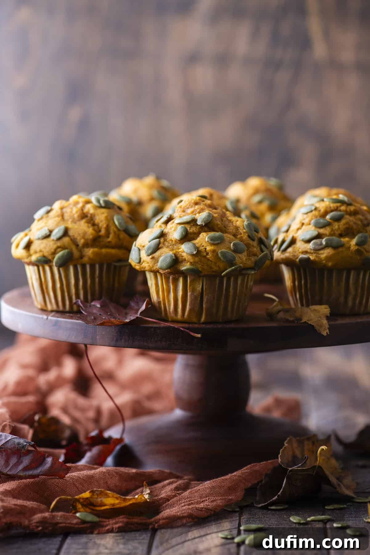 pumpkin muffins topped with pumpkin seeds on a wood cake stand surrounded by pumpkin seeds and fall leaves