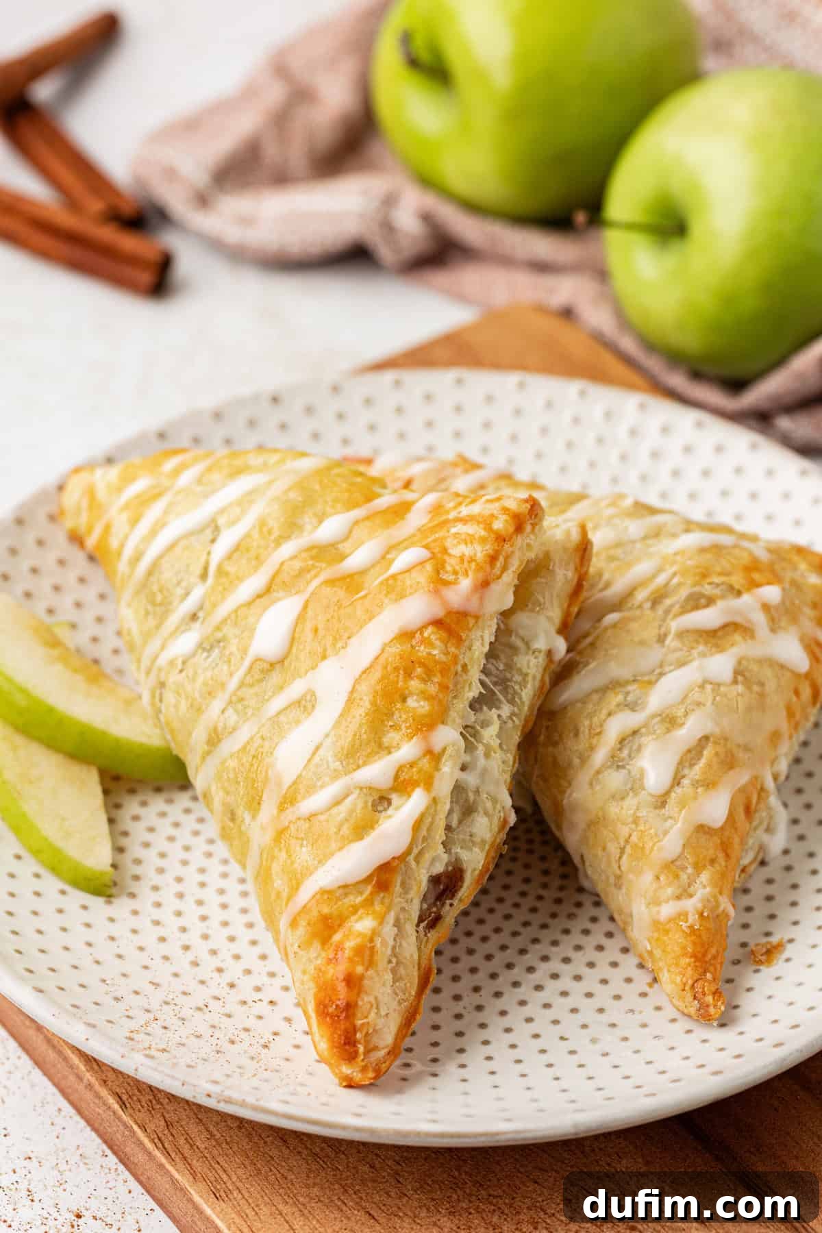 Golden Flaky Apple Turnovers 2 Two golden-brown apple turnovers on a small white plate with fresh green apple slices, whole green apples, and cinnamon sticks in the background, showcasing a rustic autumnal dessert.