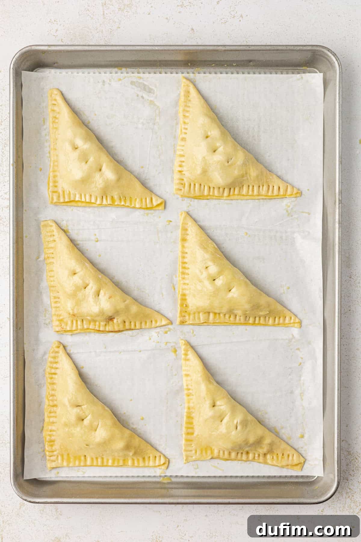 Golden Flaky Apple Turnovers 13 Six unbaked apple turnovers, perfectly formed and ready to bake, arranged in two rows on a parchment-lined baking sheet.