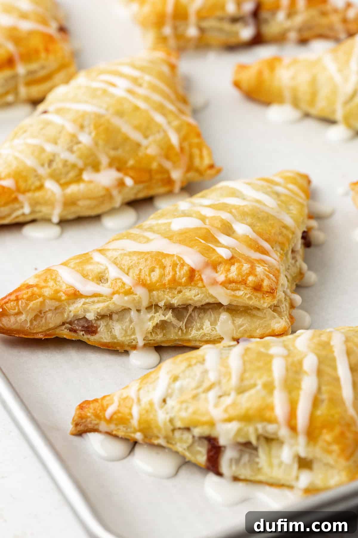 Golden Flaky Apple Turnovers 15 A close-up view of three freshly baked apple turnovers, generously drizzled with a smooth white glaze, resting on a parchment-lined baking sheet.
