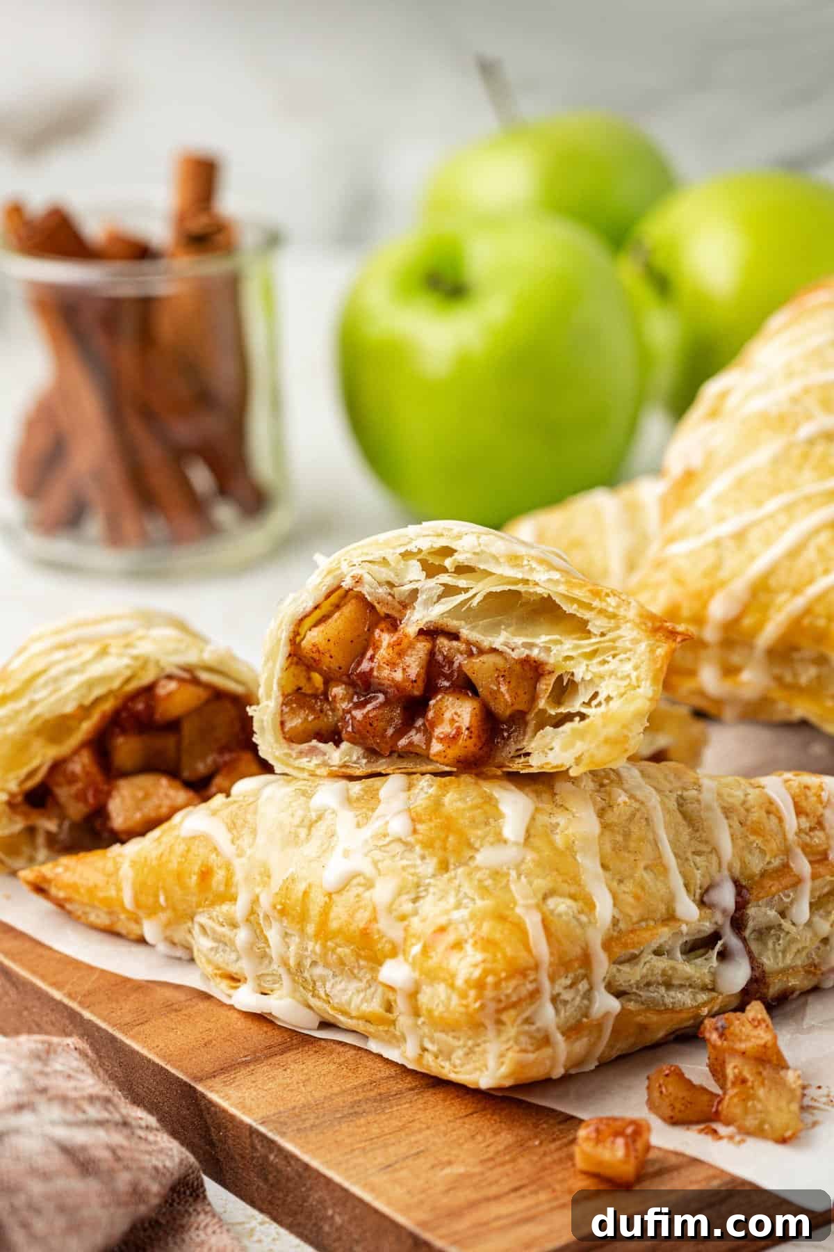 Golden Flaky Apple Turnovers 16 A perfectly baked apple turnover cut in half, revealing the soft, spiced apple filling inside. One half leans on a whole turnover, with more turnovers, fresh green apples, and cinnamon sticks blurred in the background.