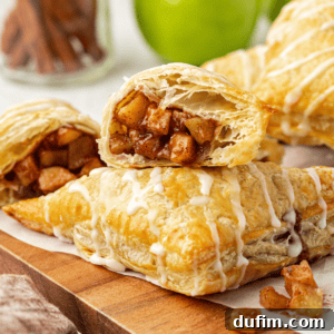 Two halves of an apple turnover, showing the moist, spiced apple filling inside. One half leans on a whole turnover, with more pastries in the blurred background.