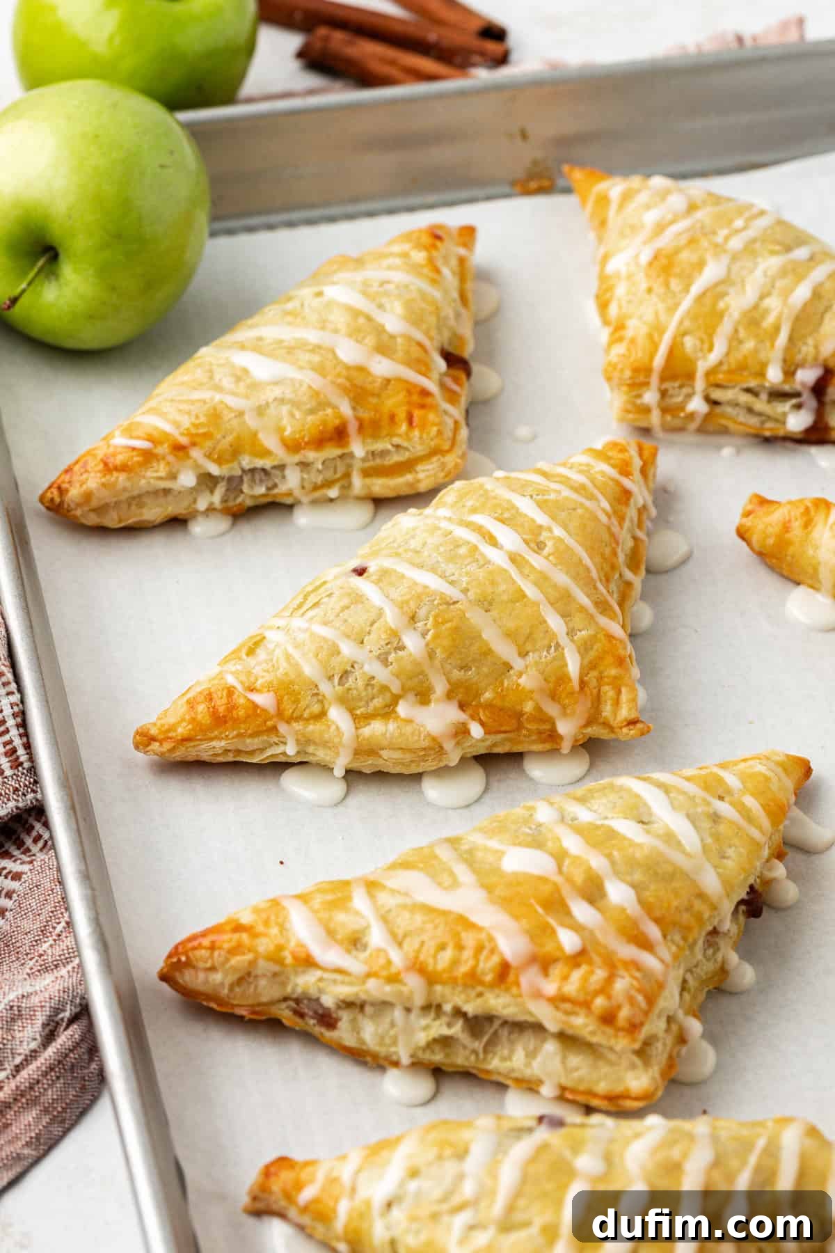 Golden Flaky Apple Turnovers 3 A row of golden-brown apple turnovers, glistening with sweet glaze, arranged on a parchment-lined baking sheet. Two whole green apples and cinnamon sticks are artfully placed around them.