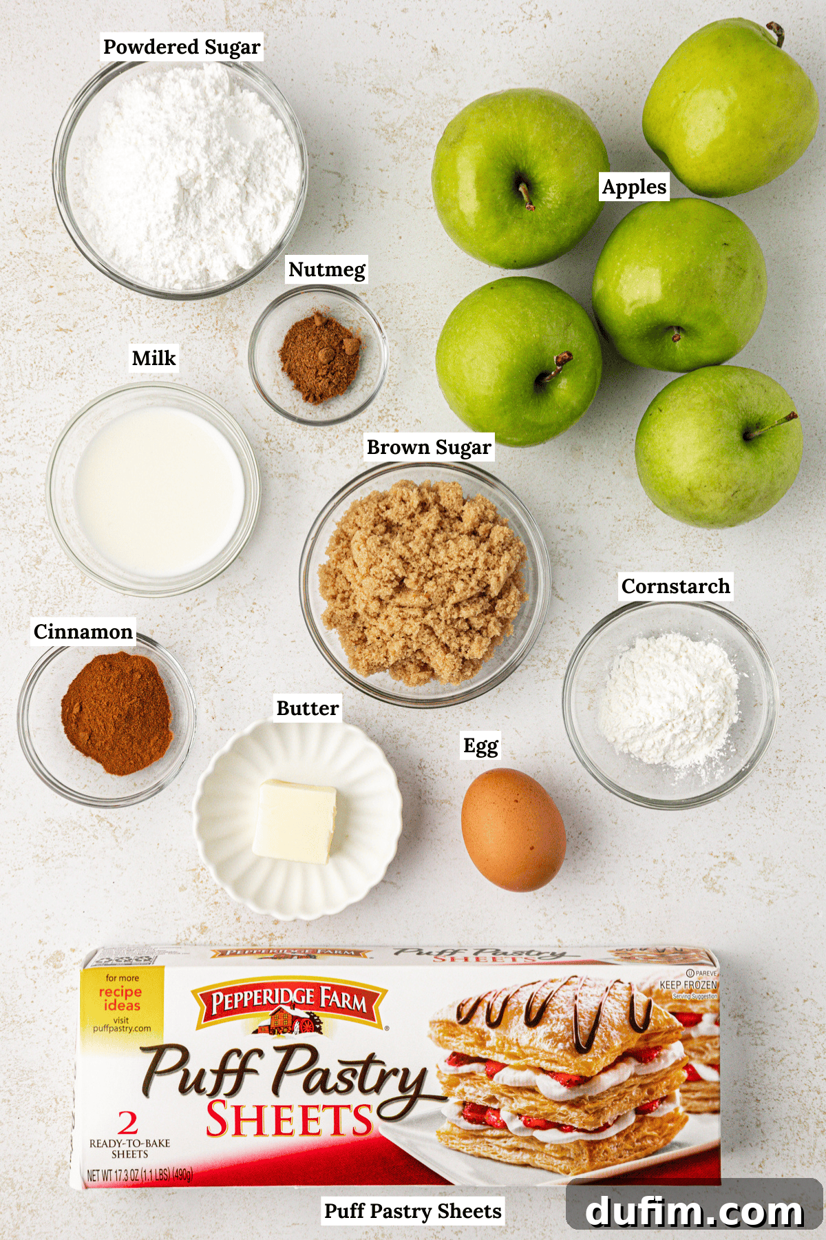 Golden Flaky Apple Turnovers 4 An overhead shot displaying all the fresh ingredients needed for apple turnovers, including powdered sugar, nutmeg, diced apples, milk, brown sugar, cinnamon sticks, butter, an egg, cornstarch, and sheets of puff pastry.