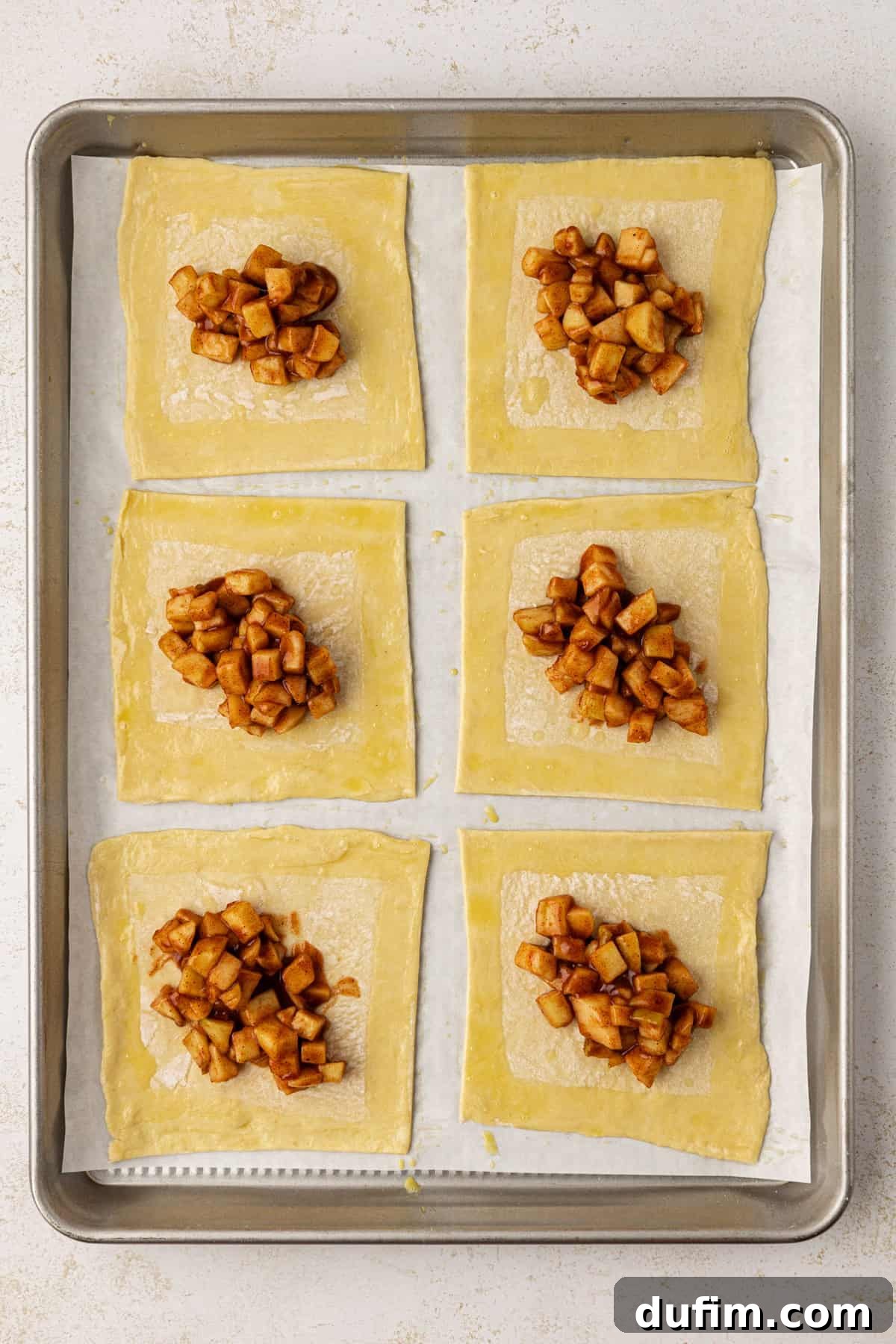 Golden Flaky Apple Turnovers 10 Six puff pastry squares, each with a dollop of apple filling in the center, neatly arranged in two rows on a parchment-lined baking sheet, prepared for folding.