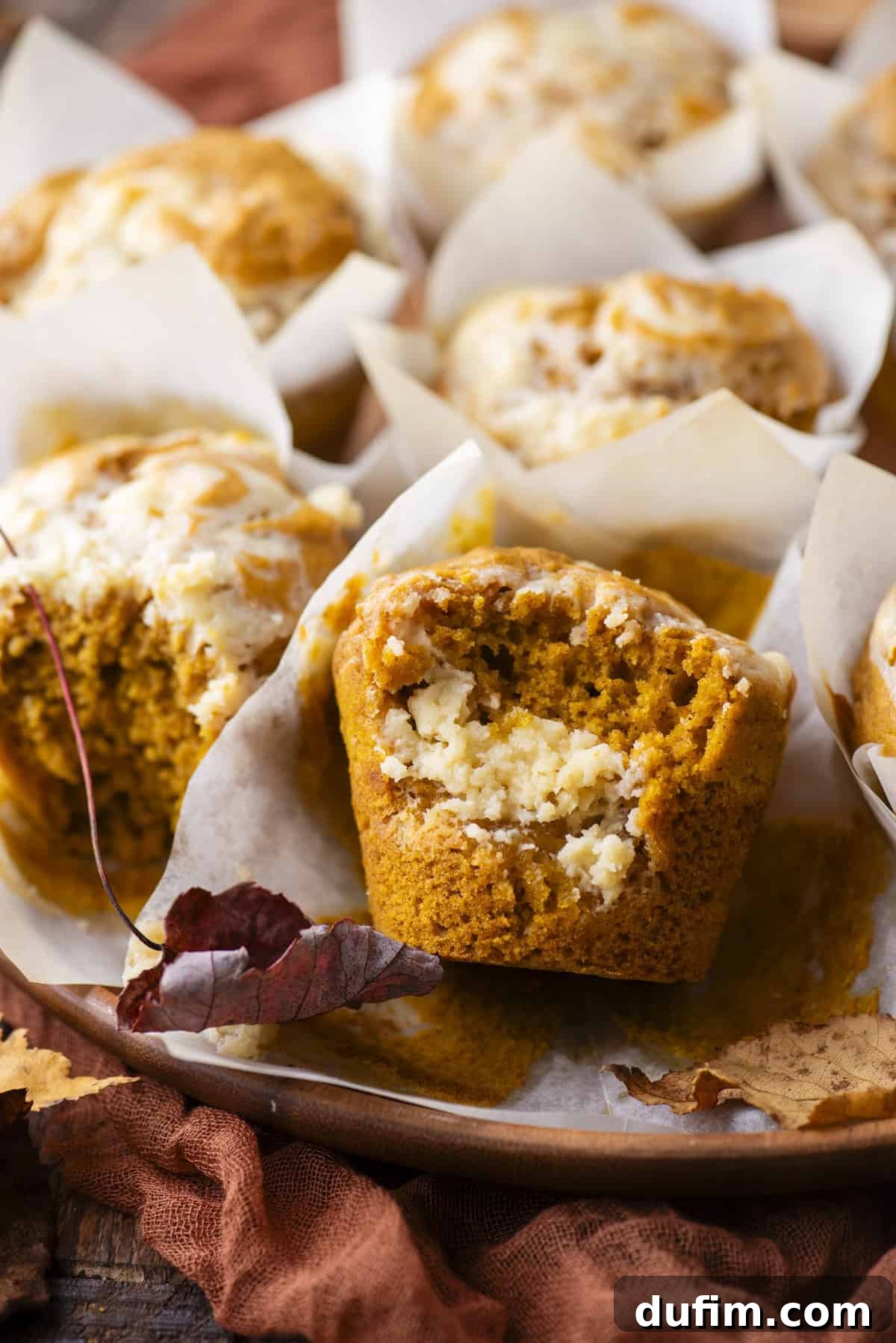 a pumpkin cream cheese muffin with a bite missing out of if, on a wood platter full of muffins surrounded by fall leaves