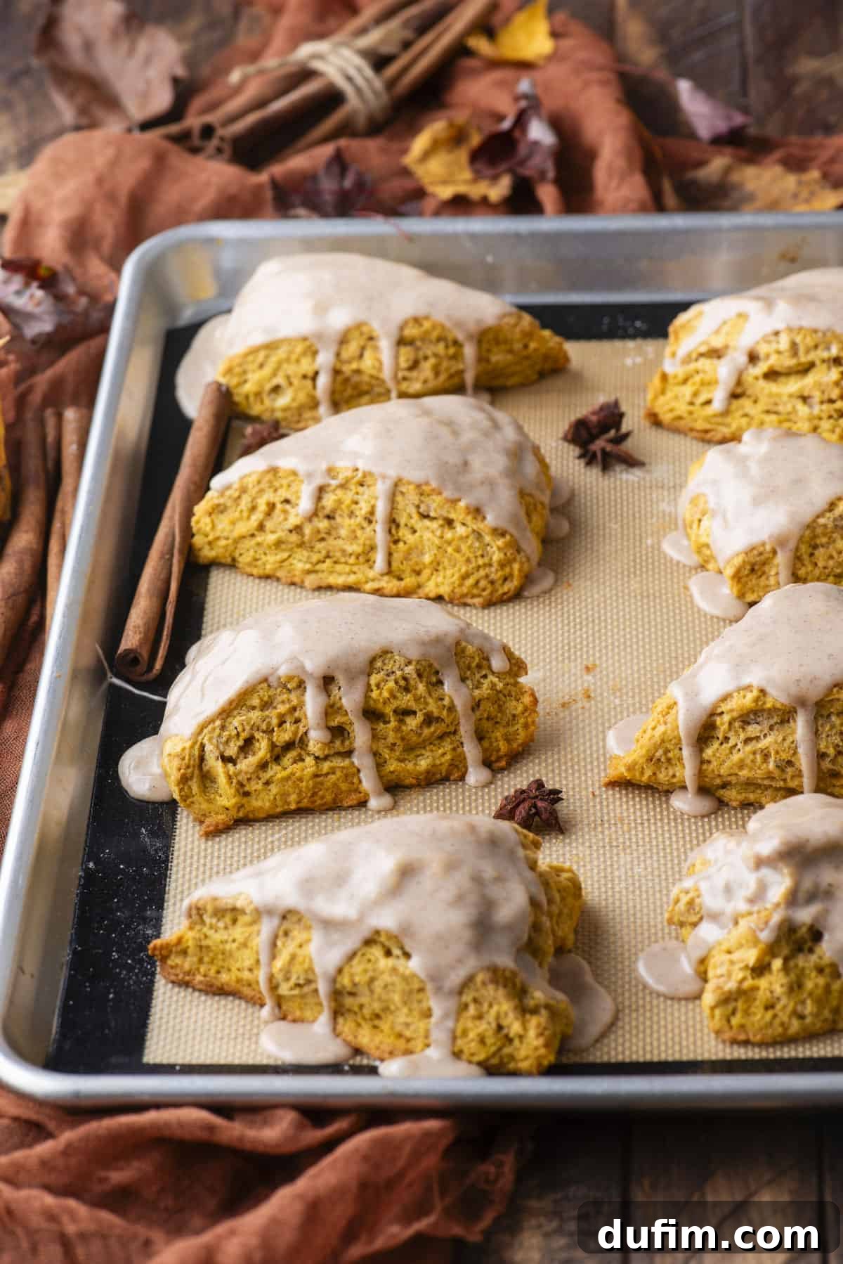 A baking sheet lined with a silicone mat, showcasing two rows of perfectly glazed pumpkin scones. Autumn leaves, cinnamon sticks, and star anise are scattered artfully around, evoking the cozy essence of fall.