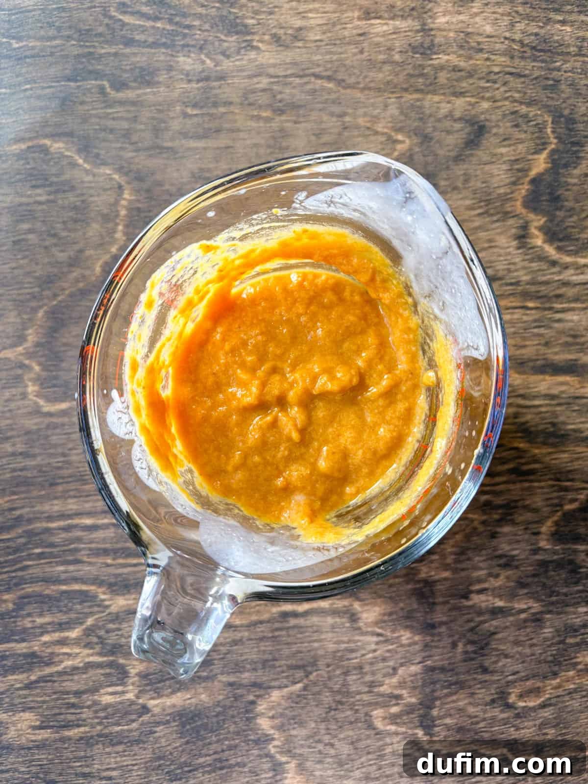 Overhead view of a large clear glass measuring cup holding a perfectly blended pumpkin puree mixture. This close-up shows the smooth, uniform consistency of the wet ingredients, ready to be incorporated into the dry flour and butter blend for the scone dough.
