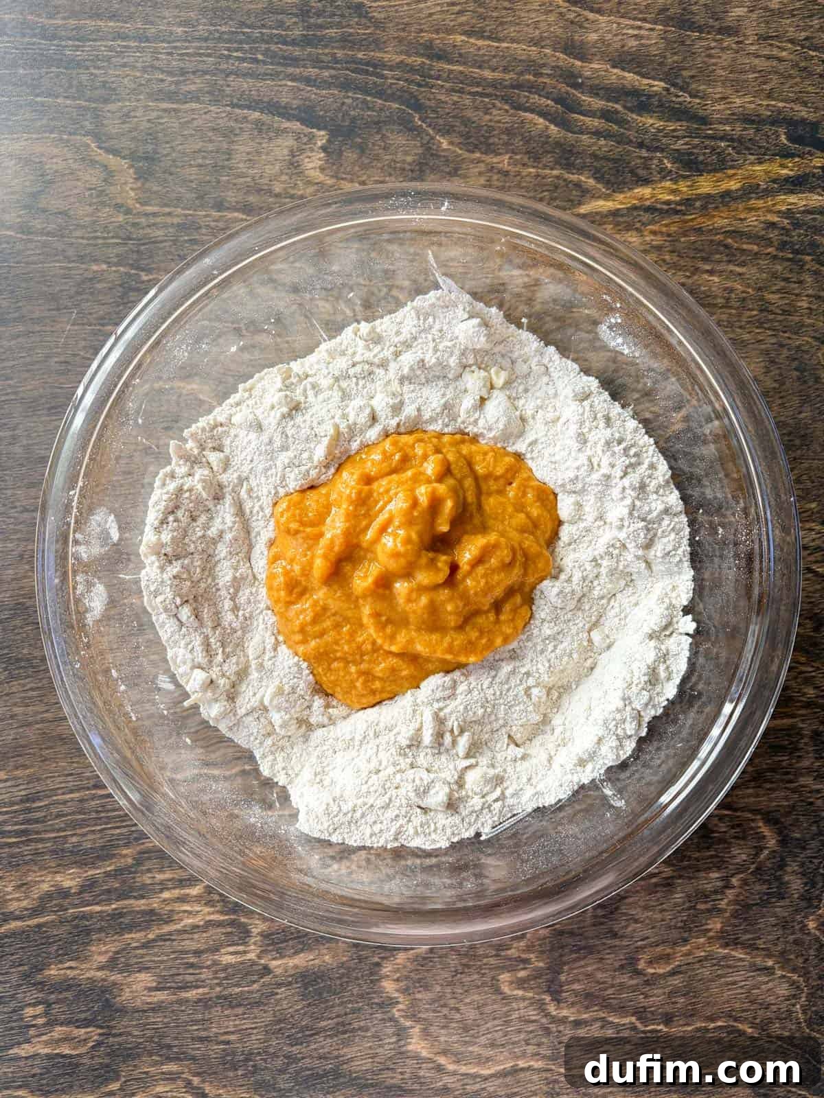 Overhead view of a clear glass bowl with a well-defined flour mixture, containing pea-sized butter pieces, forming a crater in the center. The pumpkin puree mixture is clearly visible, poured into the middle, ready to be gently combined with the dry ingredients to form the scone dough.