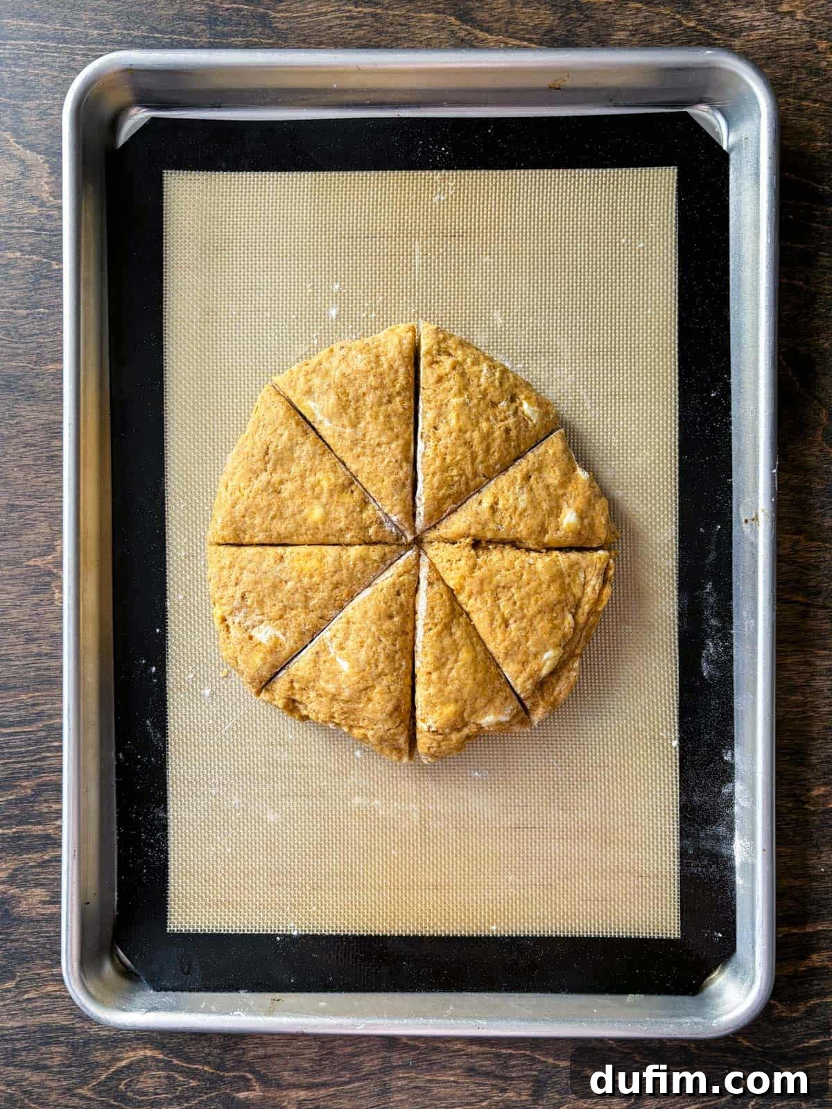 Overhead view of pumpkin scone dough, shaped into a perfect circle and precisely sliced into eight equal triangles, arranged on a baking sheet lined with a silicone mat. This preparation ensures even baking and consistent scone size.