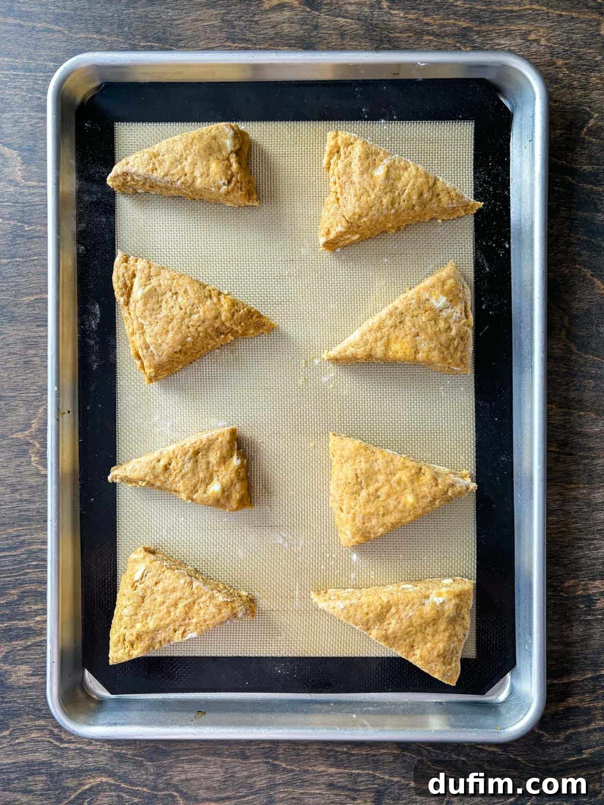 Overhead view of pumpkin scone dough, neatly cut into triangles and arranged on a baking sheet lined with a silicone baking mat, perfectly spaced and ready for the crucial freezing step before baking. The precision in cutting and spacing ensures each scone bakes evenly and rises beautifully.