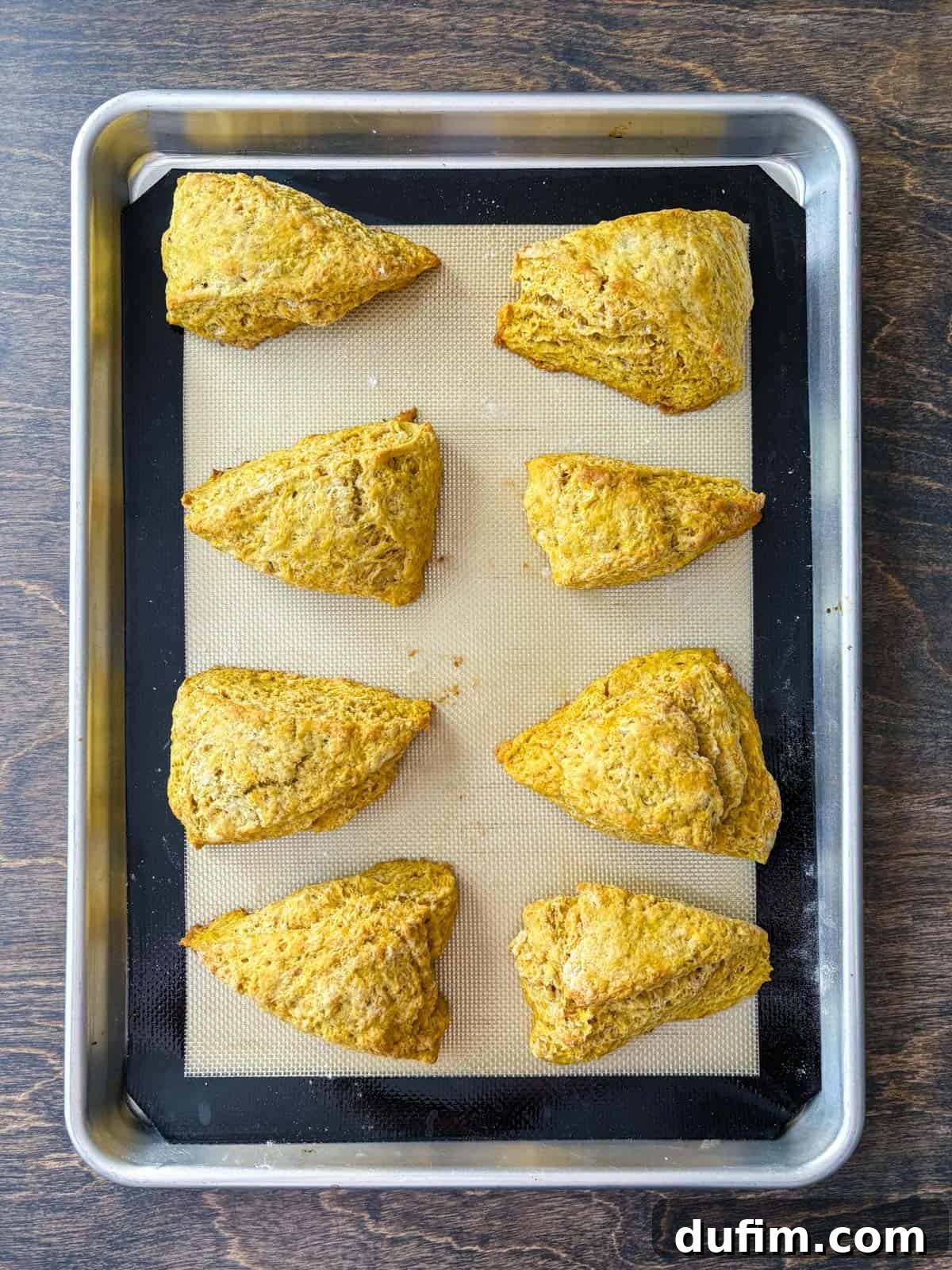 Overhead view of two neat rows of pumpkin scones, perfectly spaced on a baking sheet lined with a silicone baking mat, prepared for their final chill before baking. The uniform arrangement highlights the careful setup for consistent baking results and beautiful golden scones.