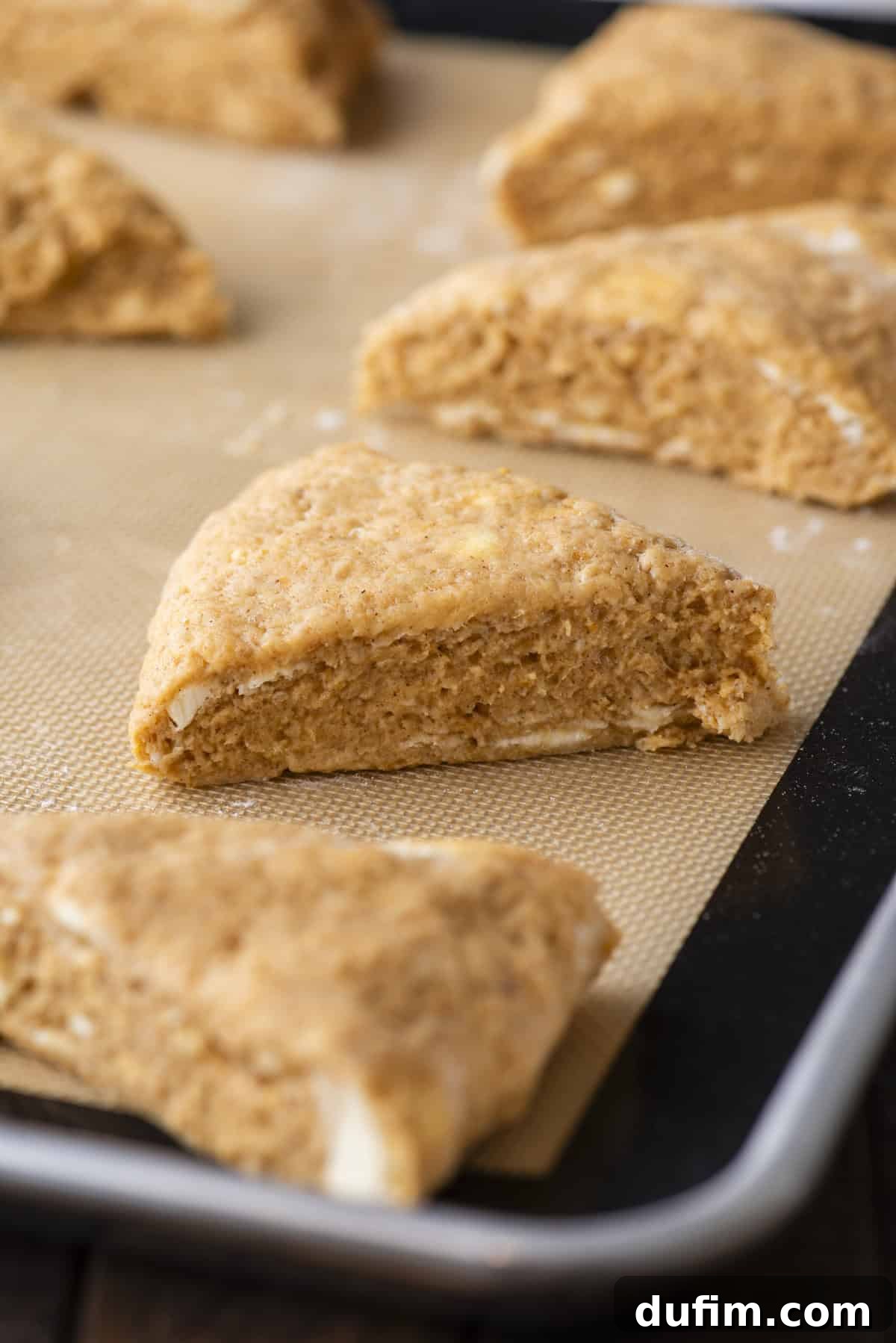 Pumpkin scone dough, neatly cut into triangles and arranged in rows on a baking sheet lined with a silicone baking mat, prepared for baking. This detailed shot shows the pre-baked scones, highlighting their uniform shape and readiness for the oven.
