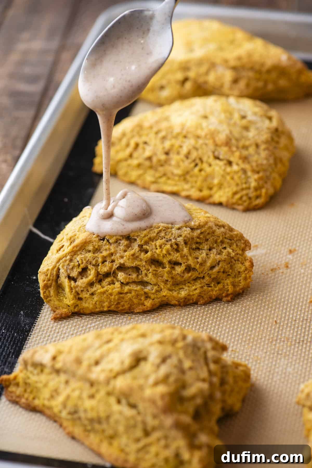 A delicate glaze being artfully drizzled over a warm pumpkin scone, creating a visually appealing finish. The scone is part of a batch arranged in rows on a baking sheet, highlighting the final decorative touch that enhances both flavor and presentation.