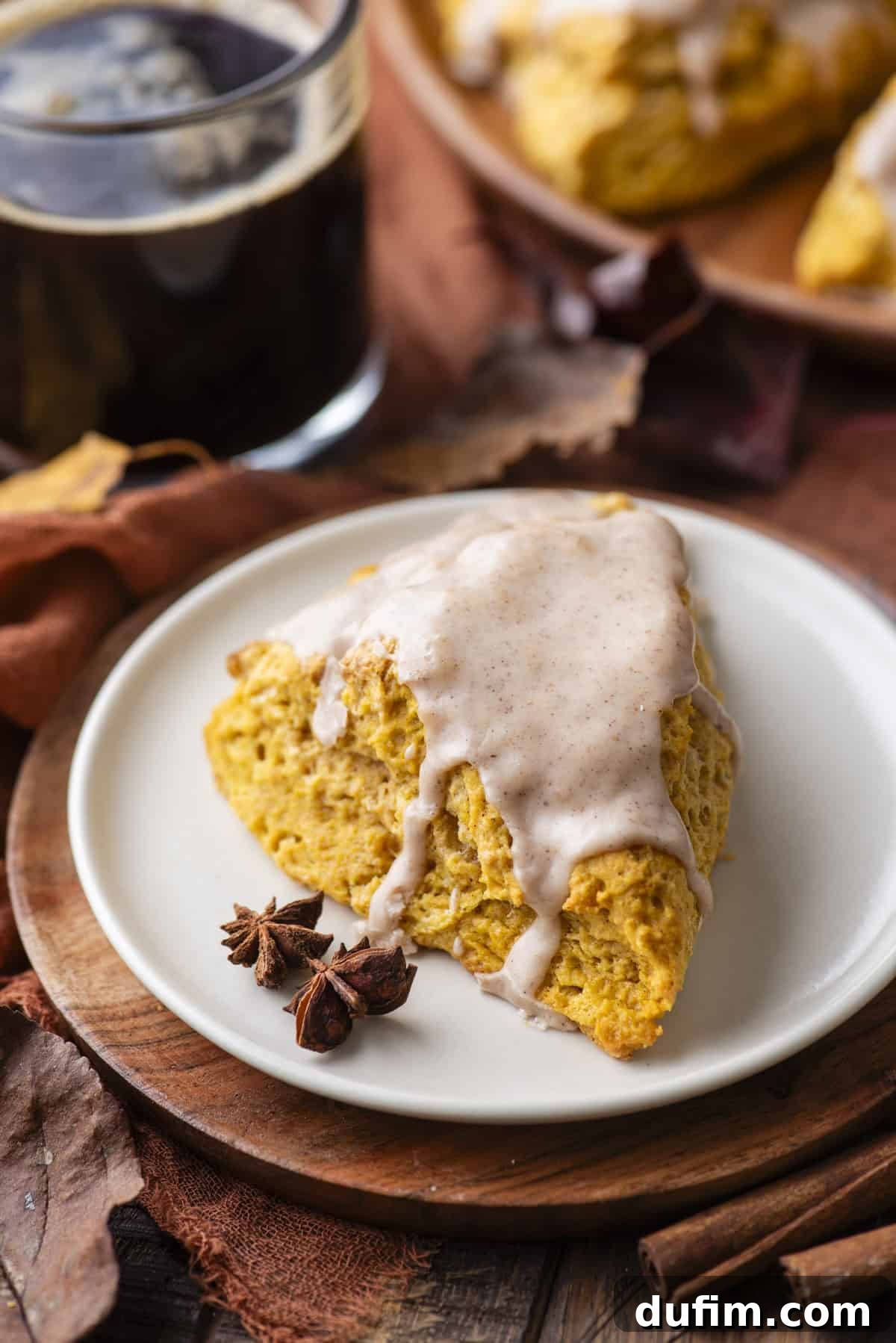 A perfectly glazed pumpkin scone, beautifully presented on a small white plate atop a rustic wood platter. A star anise pod rests beside it, while a warm cup of coffee, more scones in the background, and scattered fall leaves and cinnamon sticks complete the inviting autumn tableau.