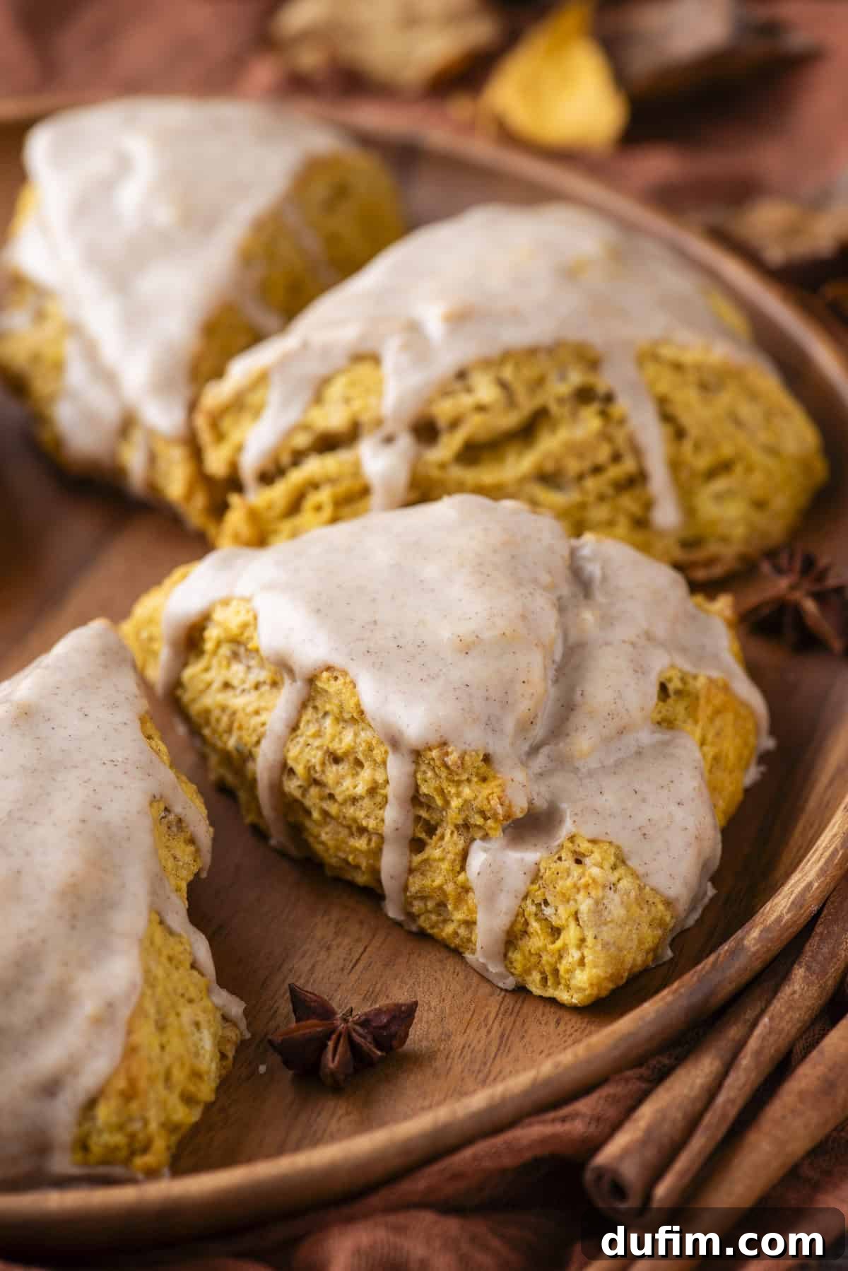 A stunning arrangement of freshly baked pumpkin scones presented on a rustic wood plate, surrounded by delicate star anise pods, vibrant autumn leaves, and fragrant cinnamon sticks. This inviting display captures the essence of fall's bounty and culinary comfort.
