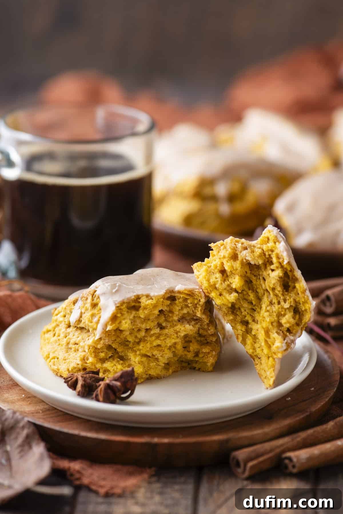 A perfectly baked pumpkin scone, artfully broken in half, with one half gently leaning against the other on a small white plate atop a rustic wood platter. A warm cup of coffee, more scones in the soft background, and scattered fall leaves, cinnamon sticks, and star anise complete the inviting autumn scene.