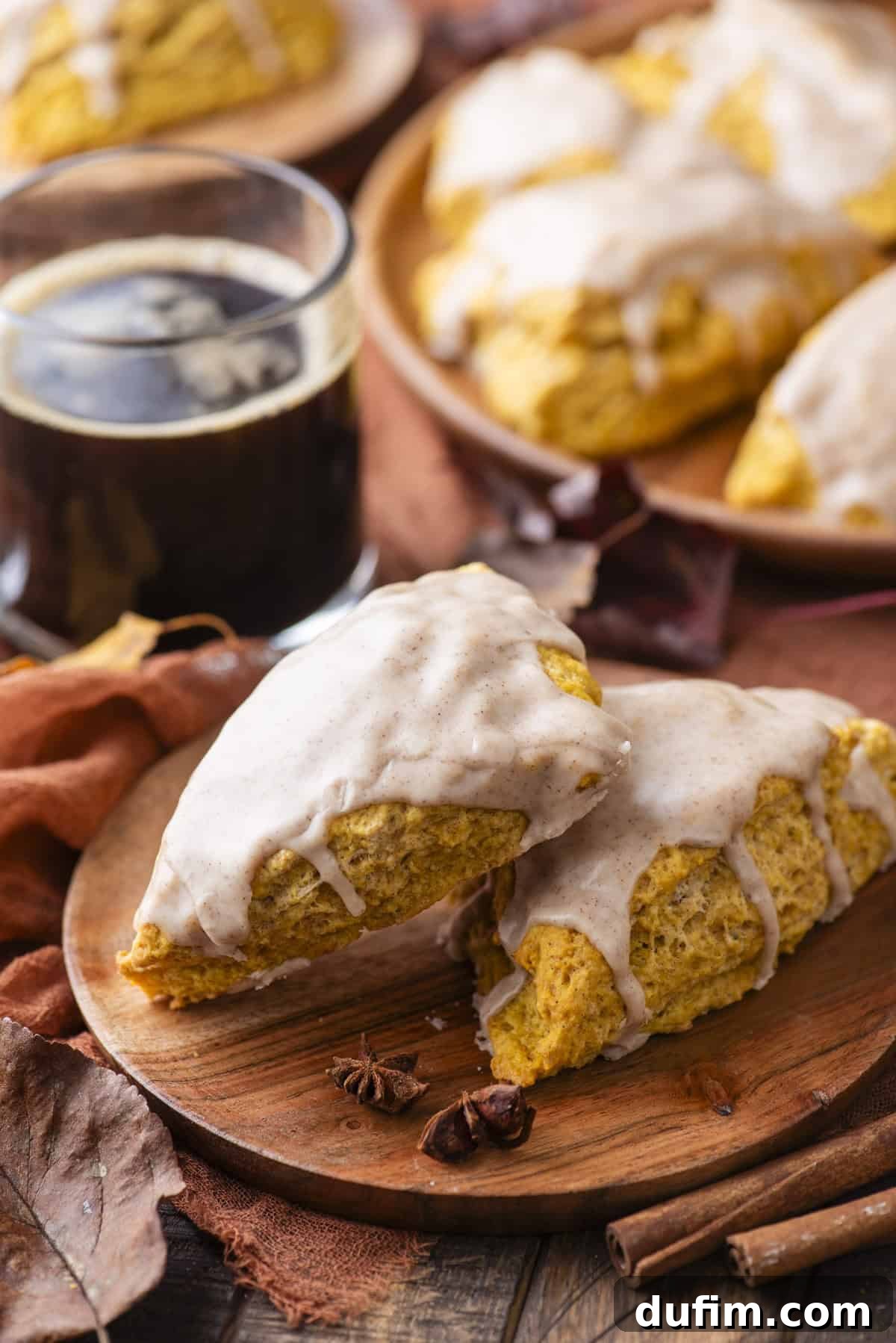 Two perfectly glazed pumpkin scones resting on a rustic wood platter, surrounded by the quintessential symbols of autumn: delicate star anise pods, vibrant fall leaves, and fragrant cinnamon sticks. A steaming cup of black coffee sits in the background, along with more scones, completing a cozy and inviting scene.