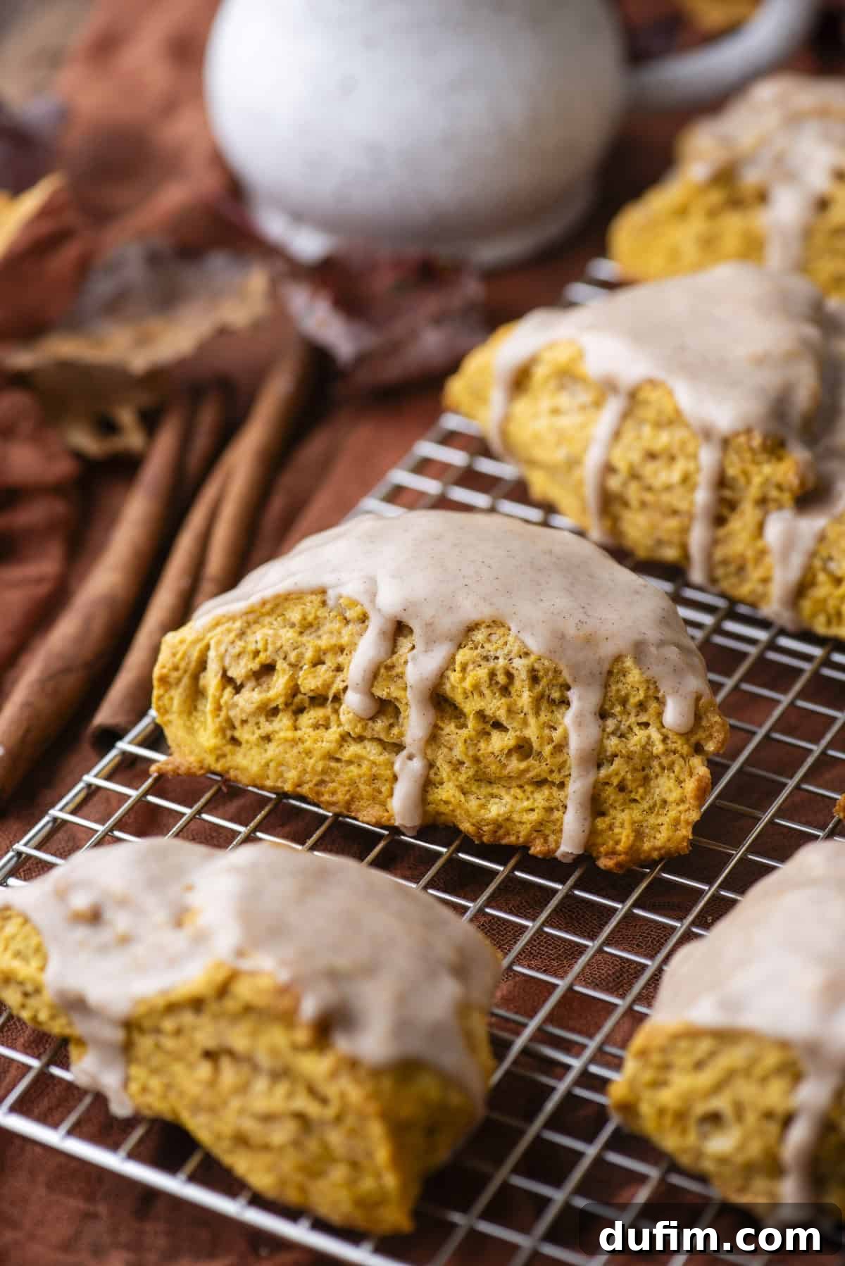 Freshly baked and glazed pumpkin scones cooling on a wire rack, with vibrant autumn leaves and fragrant cinnamon sticks scattered artfully below, creating an inviting and warm seasonal display.