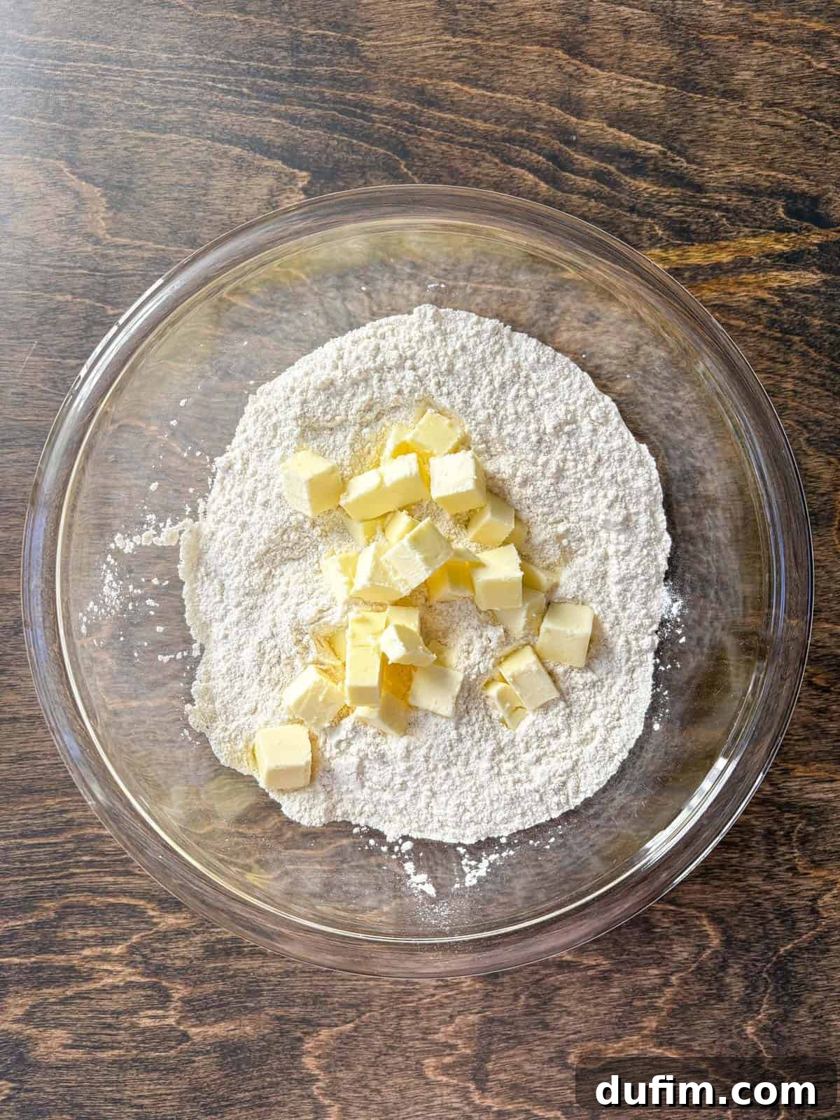 Overhead view of a clear glass bowl containing a carefully measured flour mixture, along with cubes of cold butter, ready to be incorporated for the scone dough. The dry ingredients are uniformly blended, awaiting the butter for the next crucial step in creating flaky layers.