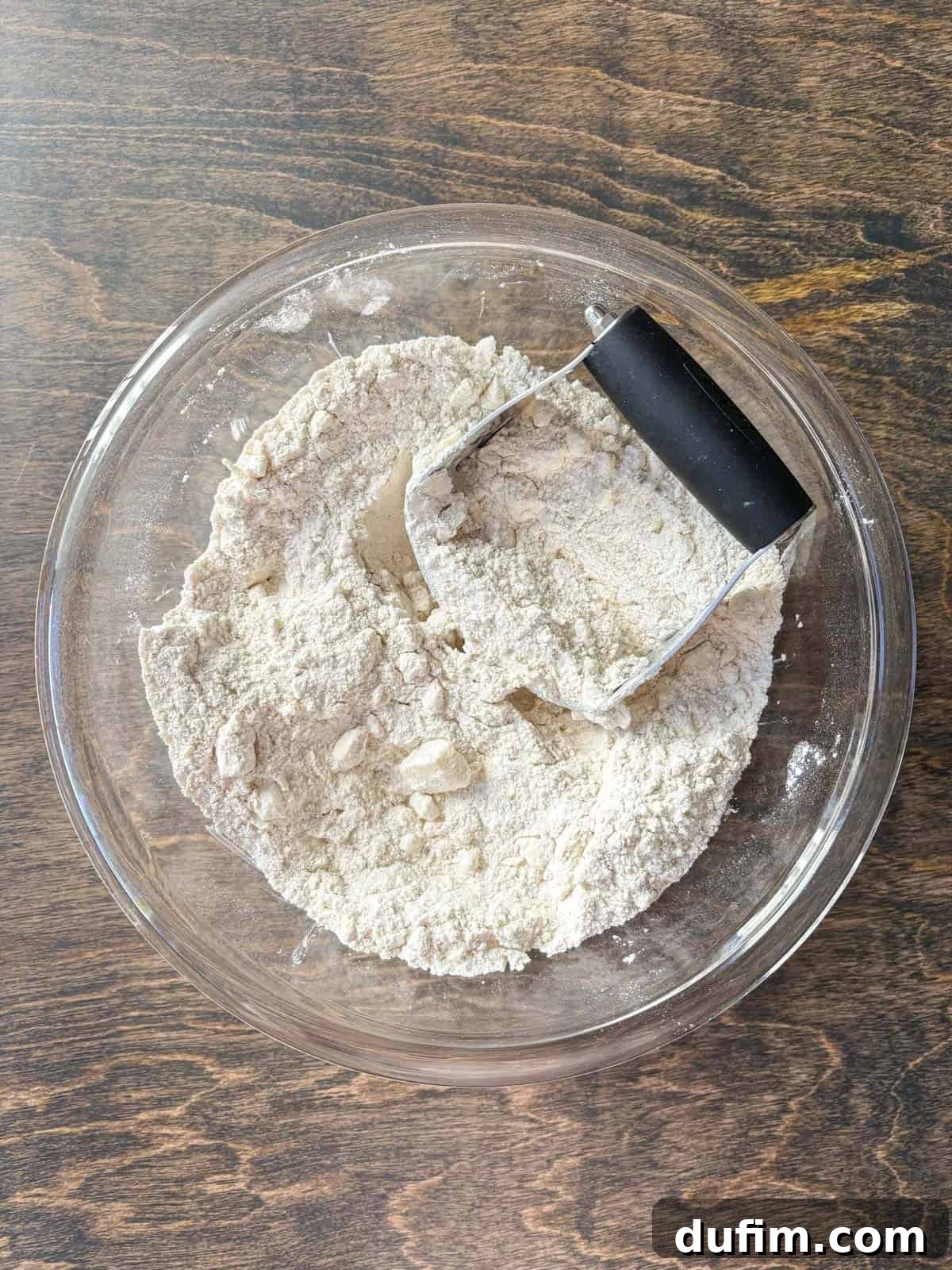 Overhead view of a clear glass bowl filled with a flour mixture and cubed cold butter, with a pastry cutter poised to begin incorporating the butter into the dry ingredients. The process aims to create pea-sized pieces of butter, essential for a light and flaky scone texture.