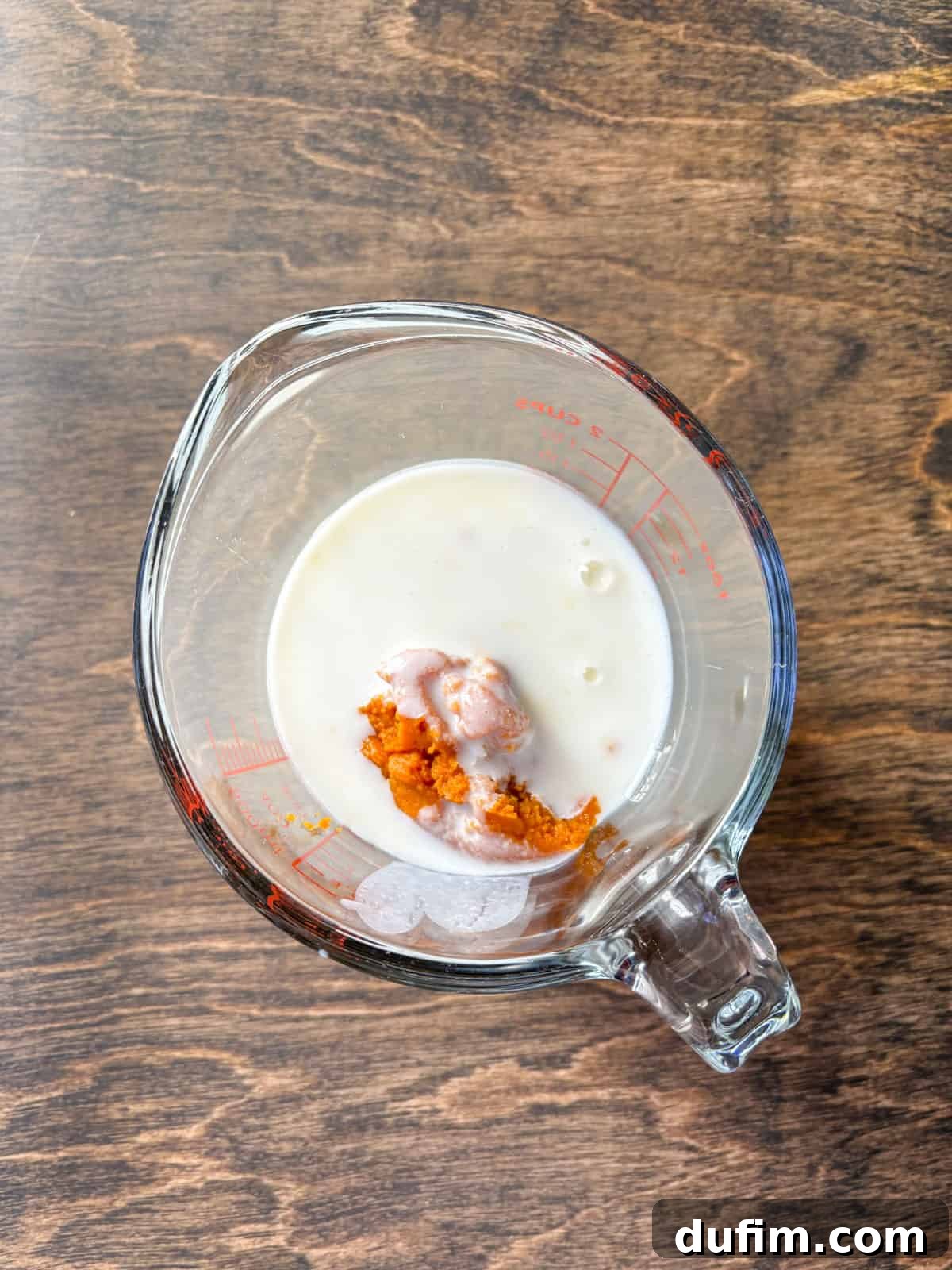 Overhead view of a large clear glass measuring cup containing creamy buttermilk and vibrant pumpkin puree, separated before mixing. This composition emphasizes the distinct liquid components that will soon be combined to form the wet ingredients for the pumpkin scones.