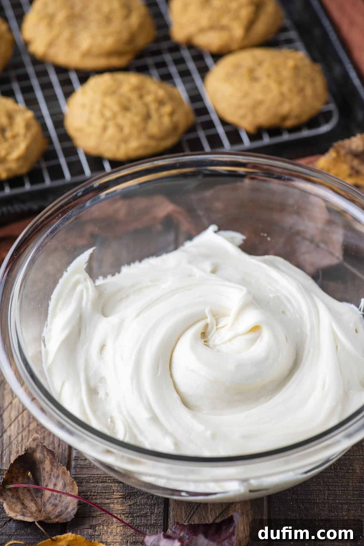 Cozy Autumn Pumpkin Spice Cookies 14 cream cheese frosting in a clear glass bowl surrounded by fall leaves beside a wire rack with pumpkin cookies in rows on it