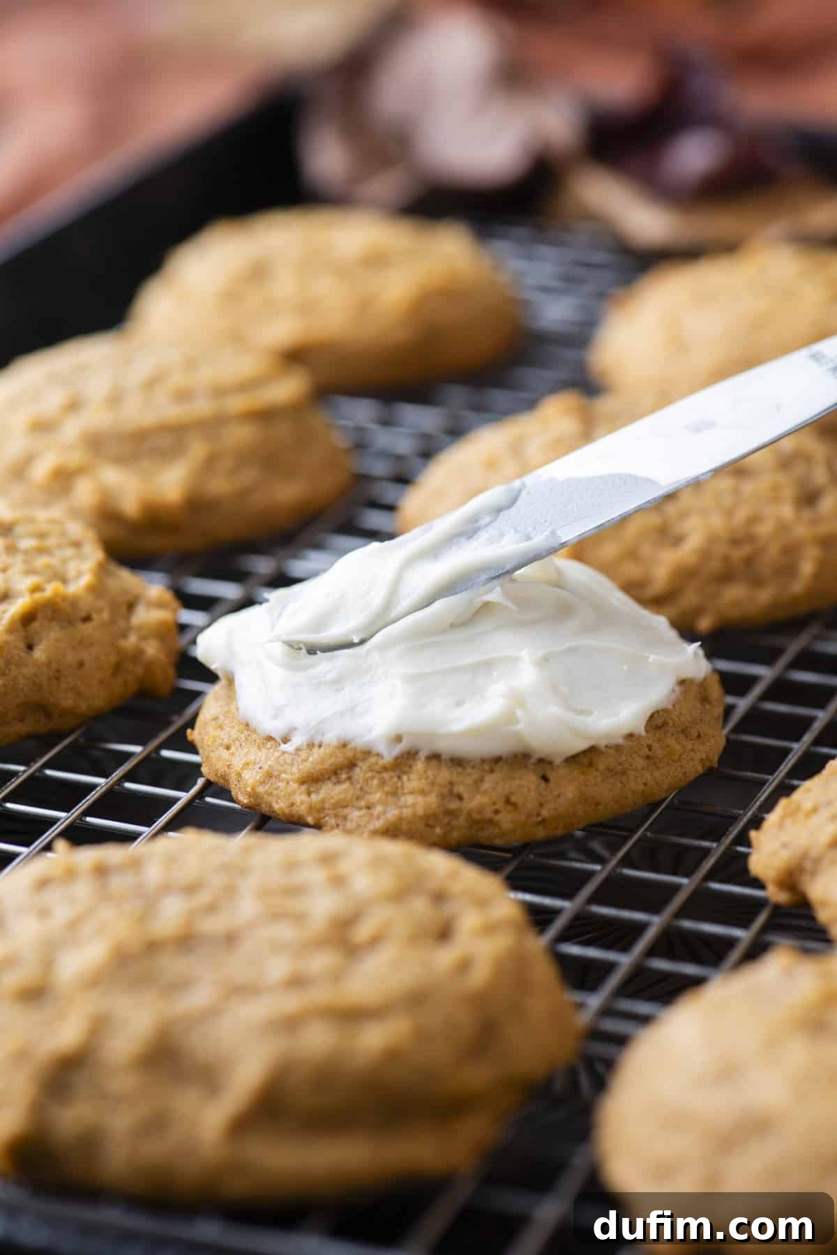 Cozy Autumn Pumpkin Spice Cookies 15 pumpkin cookies on a wire rack with one being topped with cream cheese frosting with a butter knife