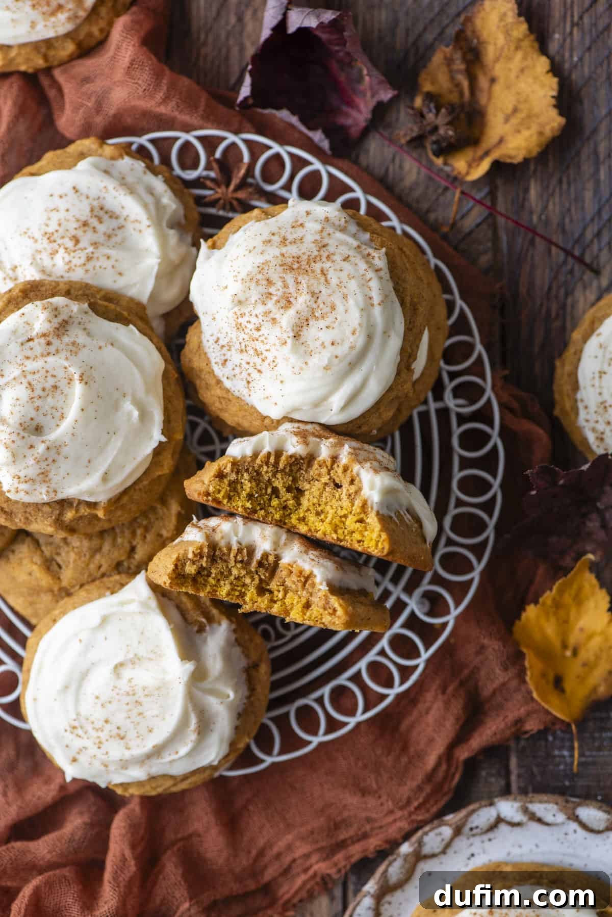 Cozy Autumn Pumpkin Spice Cookies 18 pumpkin cookies with cream cheese frosting on a white wire rack, with one cookie split in half and on its side showing the layers of cookie and frosting, surrounded by fall leaves