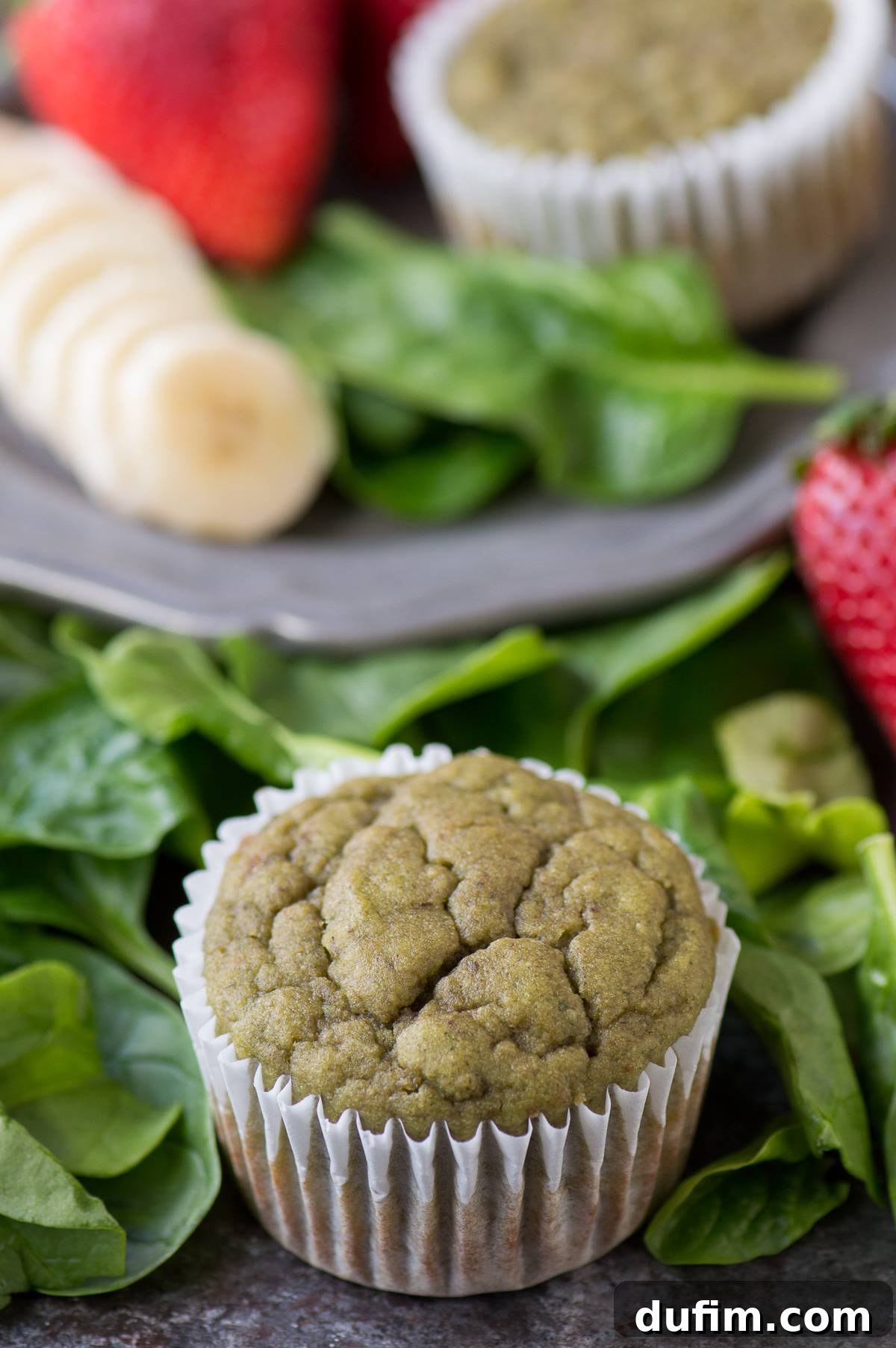 Green Monster Smoothie Muffin surrounded by fresh spinach leaves, ripe bananas, and red strawberries, showcasing its healthy ingredients.