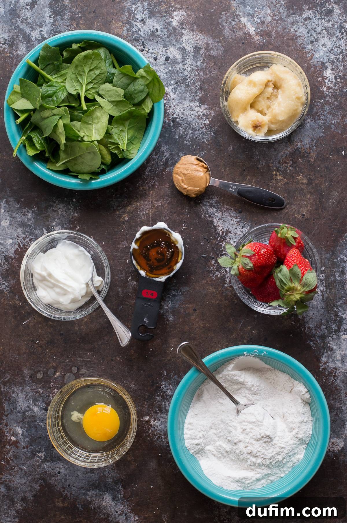 A collection of fresh ingredients laid out to make spinach muffins, including spinach leaves, bananas, strawberries, honey, peanut butter, Greek yogurt, and flour.
