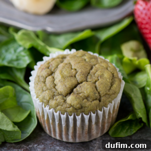 A spinach muffin in a white paper liner sits on fresh spinach leaves, with part of a strawberry and a gray plate visible in the background.