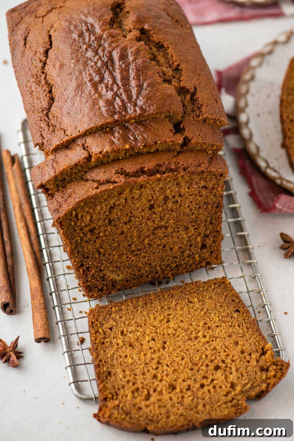 Overhead view of a golden-brown loaf of pumpkin bread on a wire cooling rack, with three inviting slices already cut. Two slices lean against the main loaf, while one lies flat, showcasing its tender crumb. Cinnamon sticks and star anise are artfully scattered around, evoking the essence of fall spices.