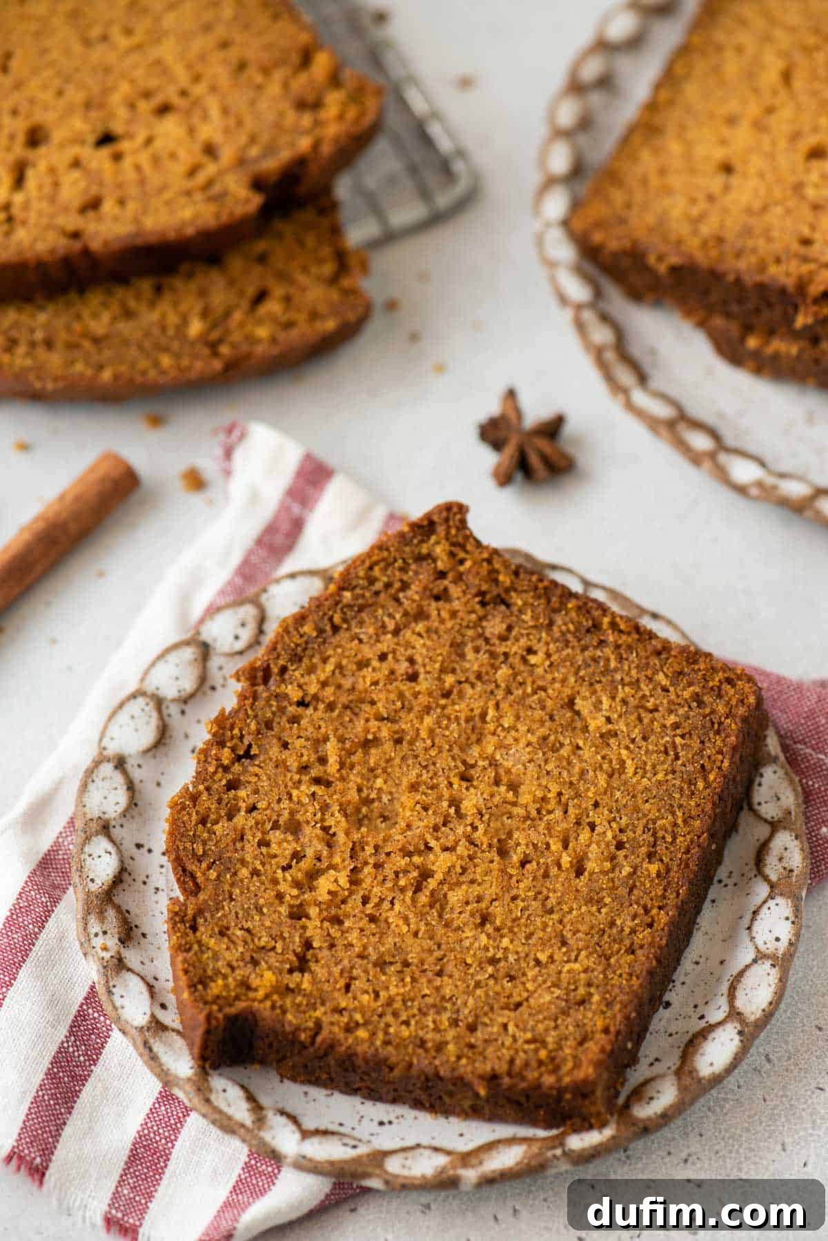 A perfectly sliced piece of pumpkin bread rests on a small, rustic plate. The plate sits atop a red and white striped towel, flanked by cinnamon sticks, star anise, and additional slices of the delicious bread, highlighting its autumnal appeal.