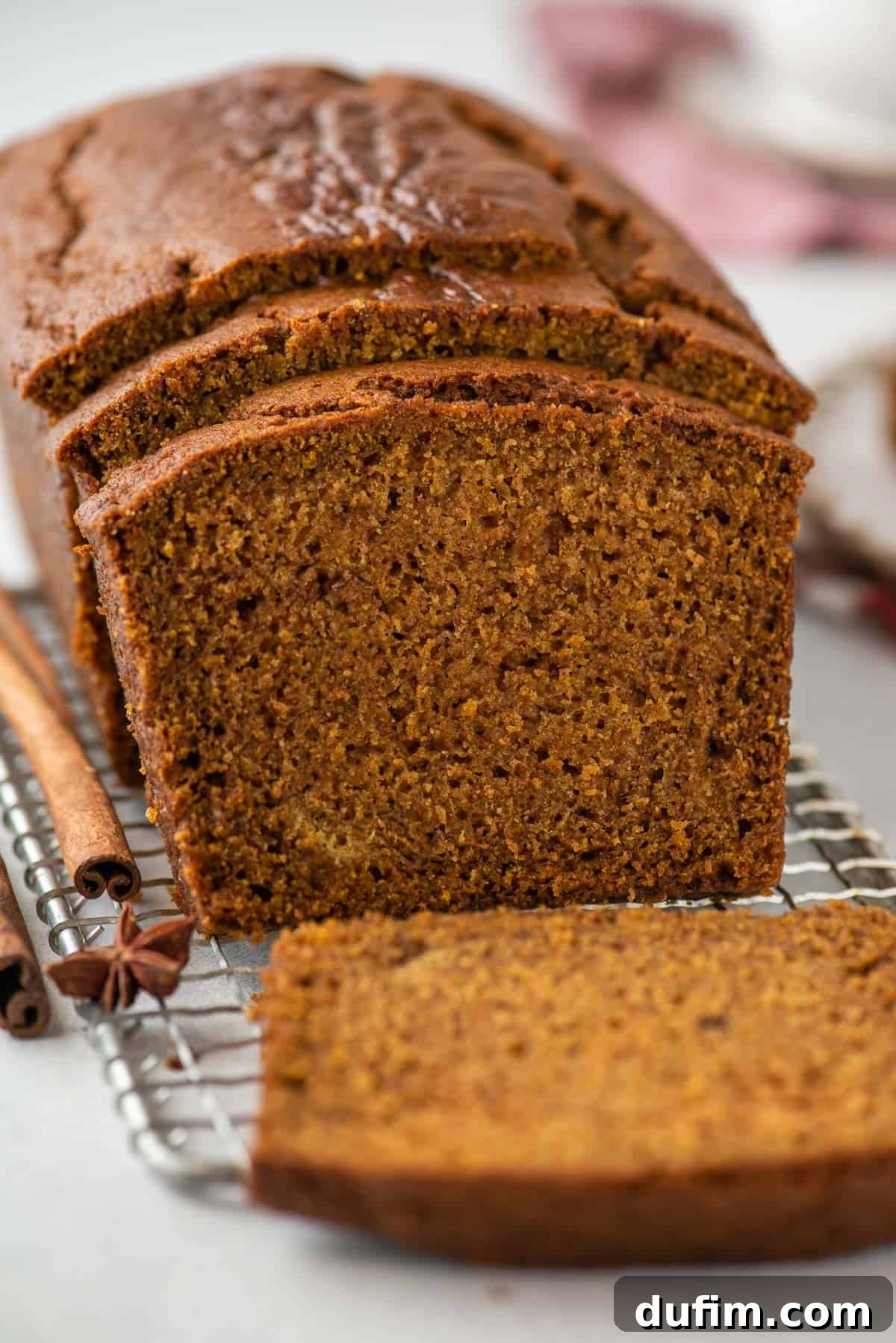 A rustic loaf of pumpkin bread, fresh from the oven, cooling on a wire rack. A single slice has been cut and laid next to the loaf, while two more lean gently against the main body of the bread. Cinnamon sticks and star anise are casually arranged around the bread, emphasizing its aromatic spices.