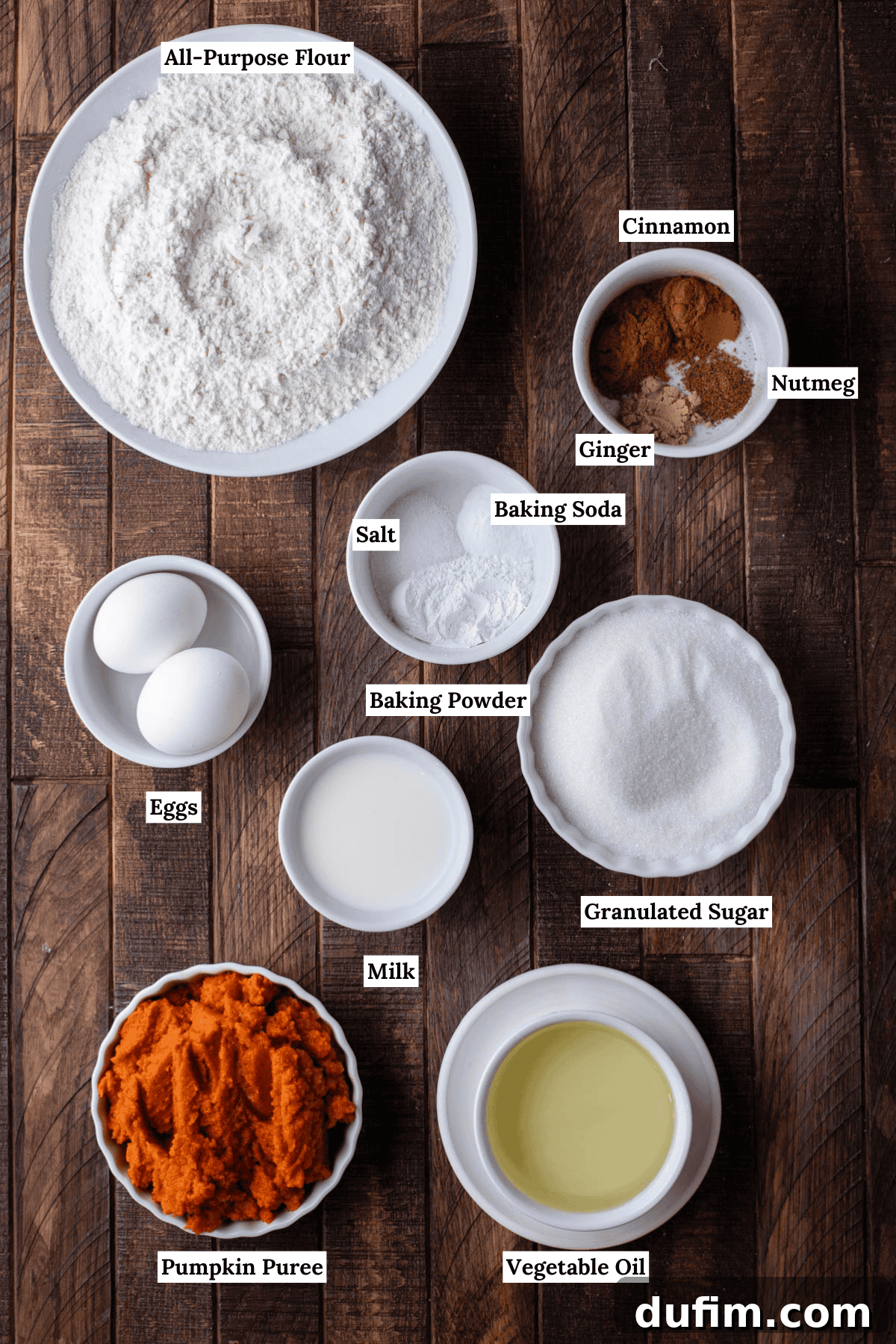 An overhead shot showcasing the neatly arranged ingredients for homemade pumpkin bread. Key components include all-purpose flour, baking powder, baking soda, salt, aromatic ground cinnamon, nutmeg, ginger, farm-fresh eggs, pure granulated sugar, milk, creamy pumpkin puree, and vegetable oil.