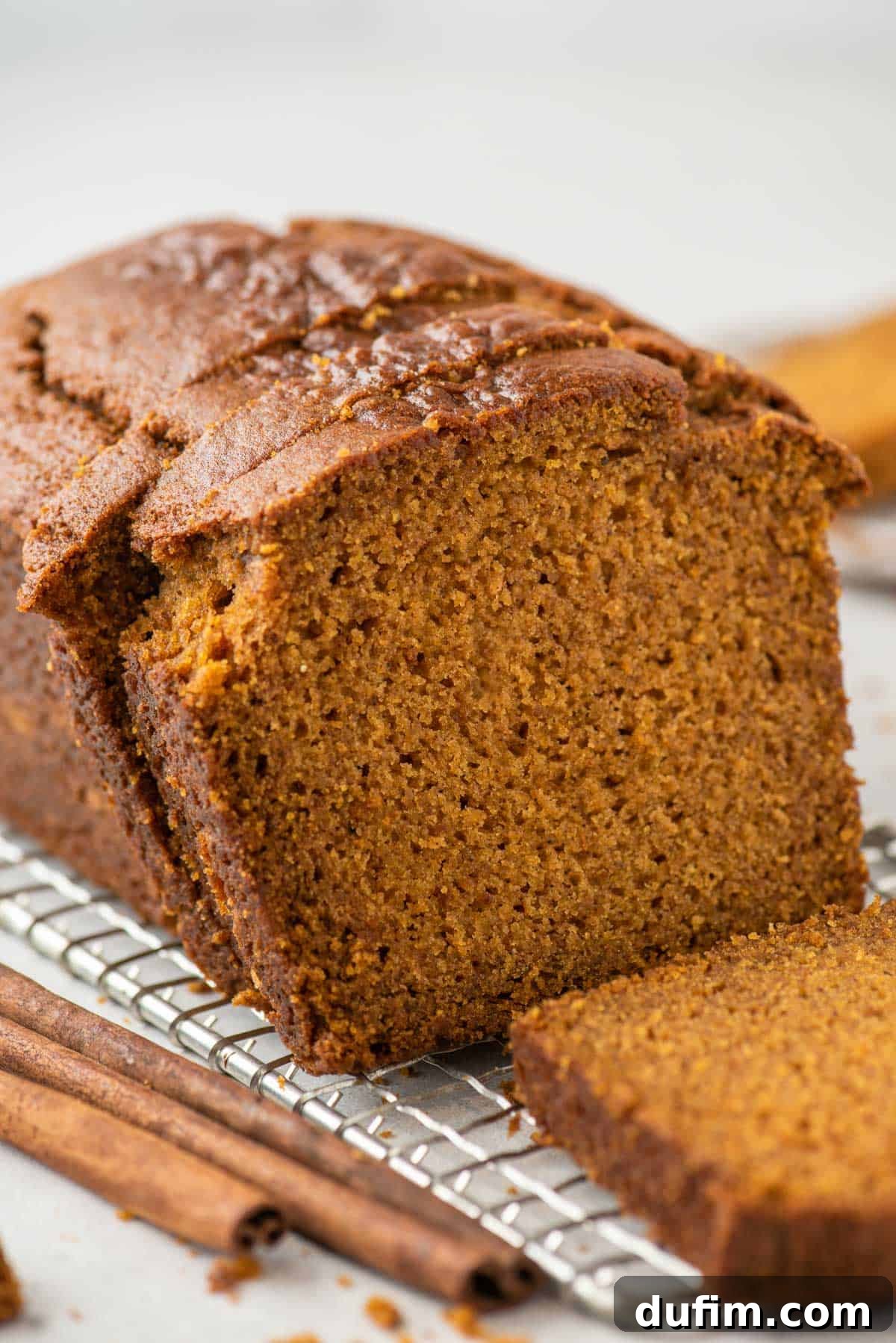 A warm loaf of pumpkin bread rests on a wire cooling rack. Two slices are gently cut and placed next to the main loaf, with one lying flat and another leaning. Three cinnamon sticks lie invitingly beside the bread, adding to the autumnal scene.