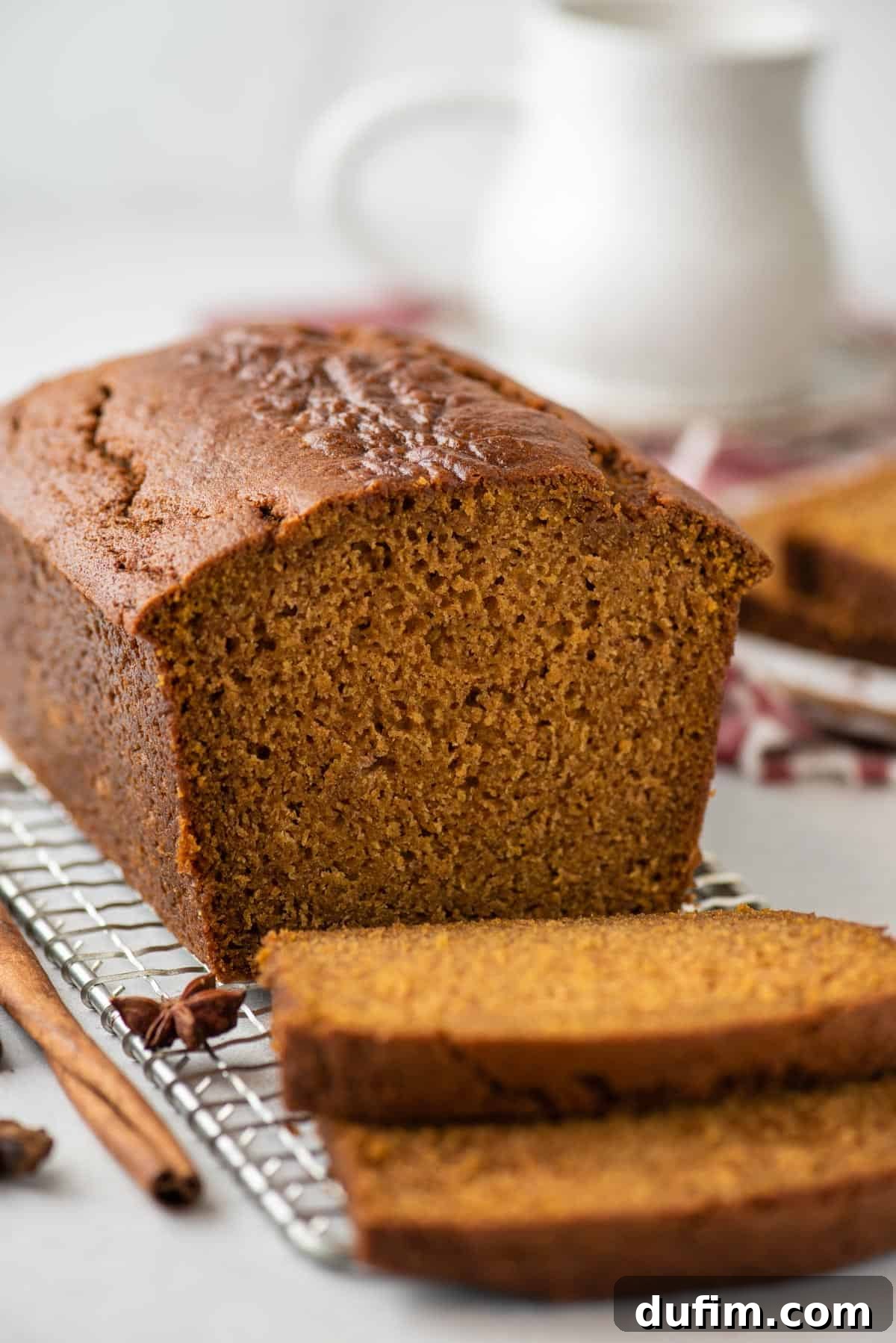 A freshly baked loaf of pumpkin bread rests on a wire cooling rack. Two slices have been cut from the loaf, one lying flat and the other leaning against it. A cinnamon stick and a star anise are gently scattered around, adding a touch of rustic charm to the scene.