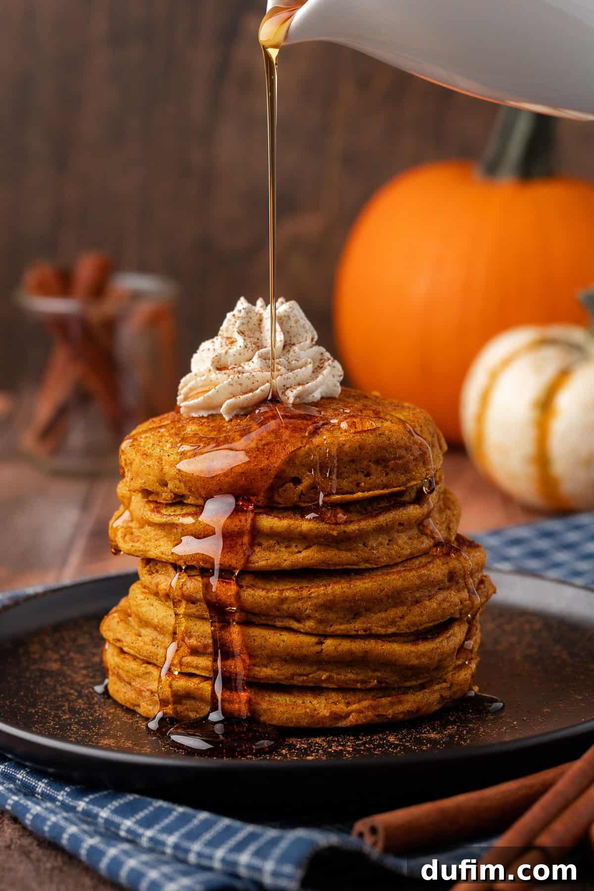 A luxurious stack of pumpkin pancakes on a black plate, generously topped with whipped cream and being drizzled with warm maple syrup, creating an inviting visual of a perfect fall breakfast.