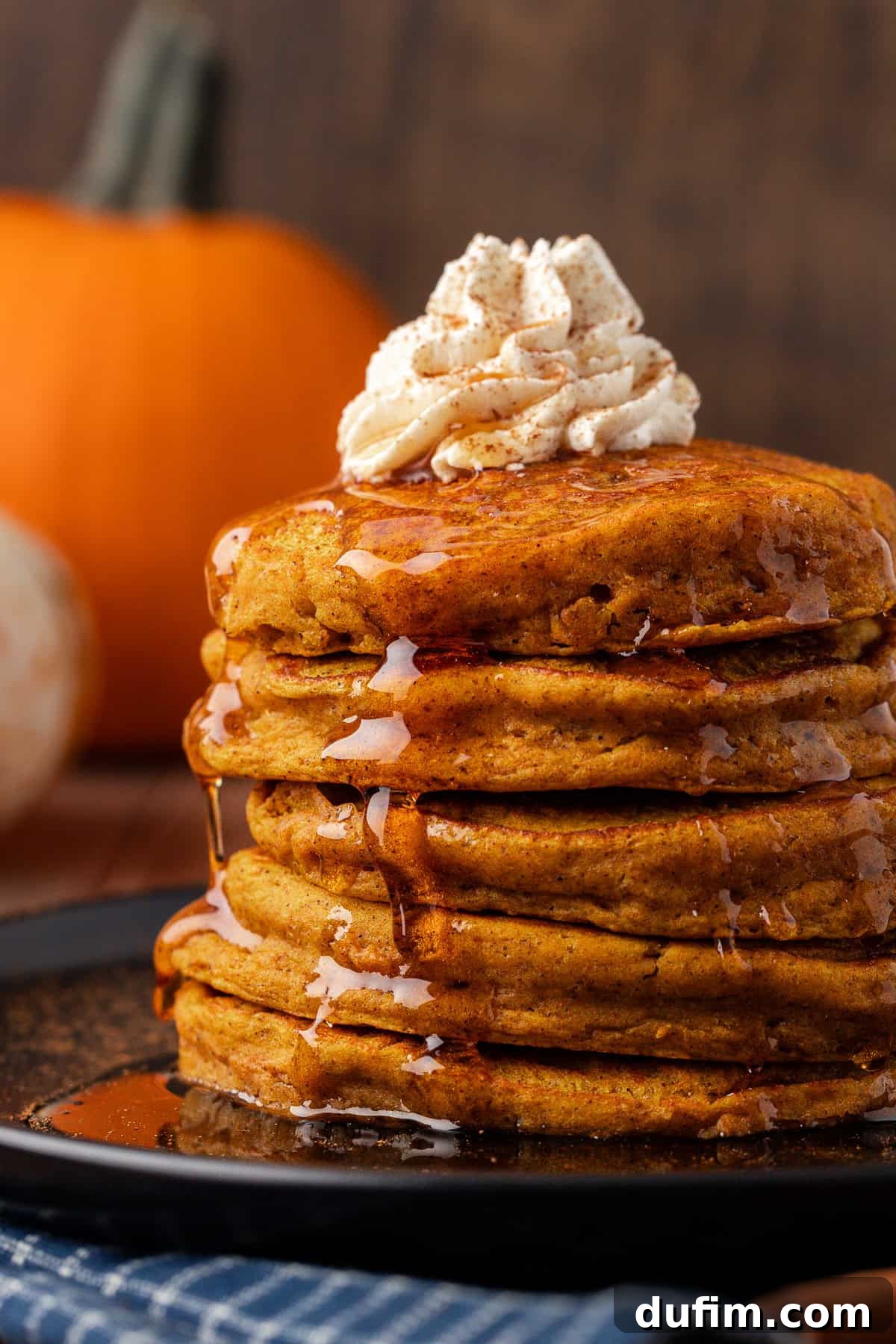 A magnificent stack of pumpkin pancakes on a black plate, crowned with fluffy whipped cream and glistening with poured syrup, ready to be devoured.