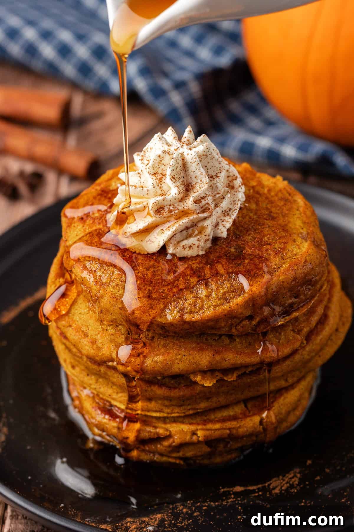 A mouthwatering stack of pumpkin pancakes on a black plate, generously topped with whipped cream and drizzled with glistening maple syrup, a true autumnal delight.