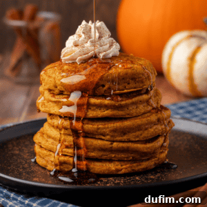 A stack of pumpkin pancakes on a black plate, topped with whipped cream, being drizzled with syrup