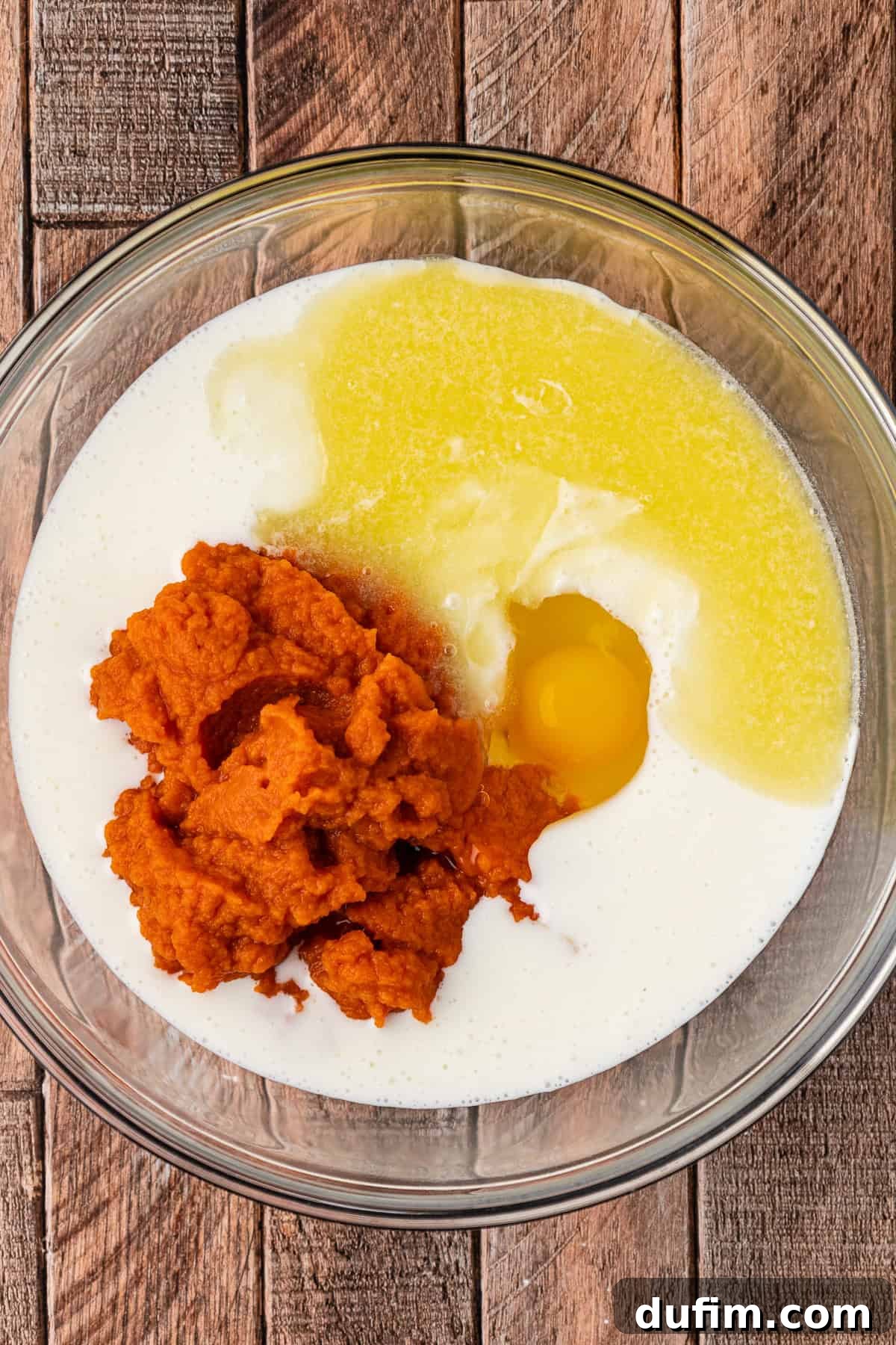 A large clear glass bowl holds the wet ingredients for pumpkin pancakes: buttermilk, pumpkin puree, and an egg, ready to be whisked together.
