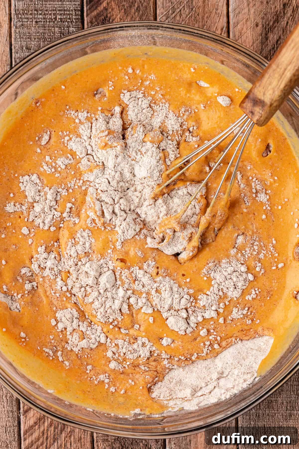 Pumpkin pancake batter being gently mixed in a large glass bowl with a whisk, showing a few lumps which are perfectly normal.