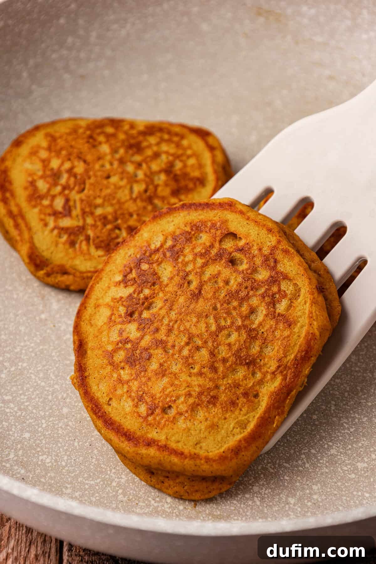 Two pumpkin pancakes cooking in a pan, with one being expertly flipped by a spatula, showing its golden-brown underside.