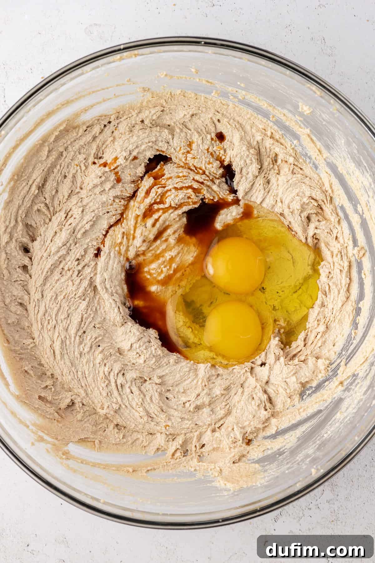 White chocolate chip cookie dough ingredients in a clear glass bowl, with egg and vanilla sitting on top not yet combined