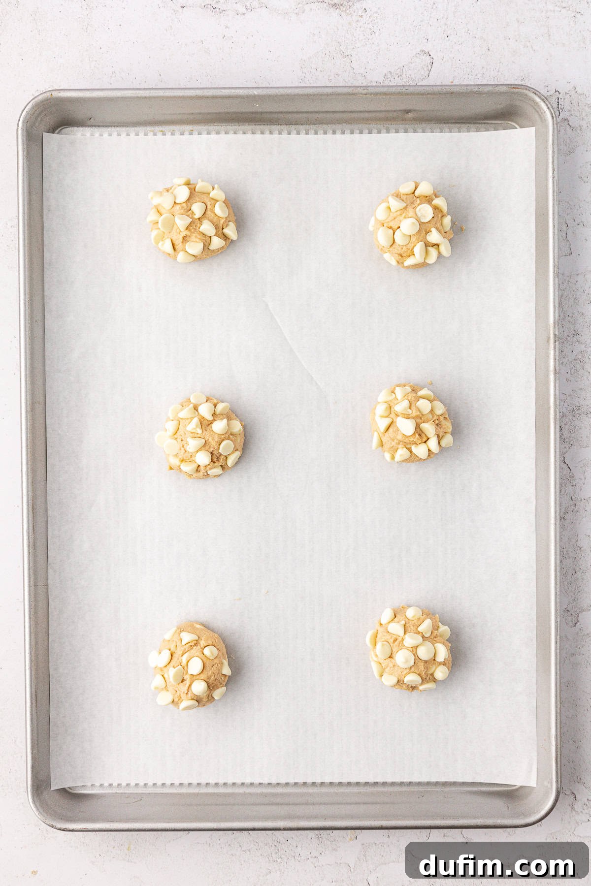Two rows of three white chocolate chip cookie dough balls on a baking sheet lined with white parchment paper