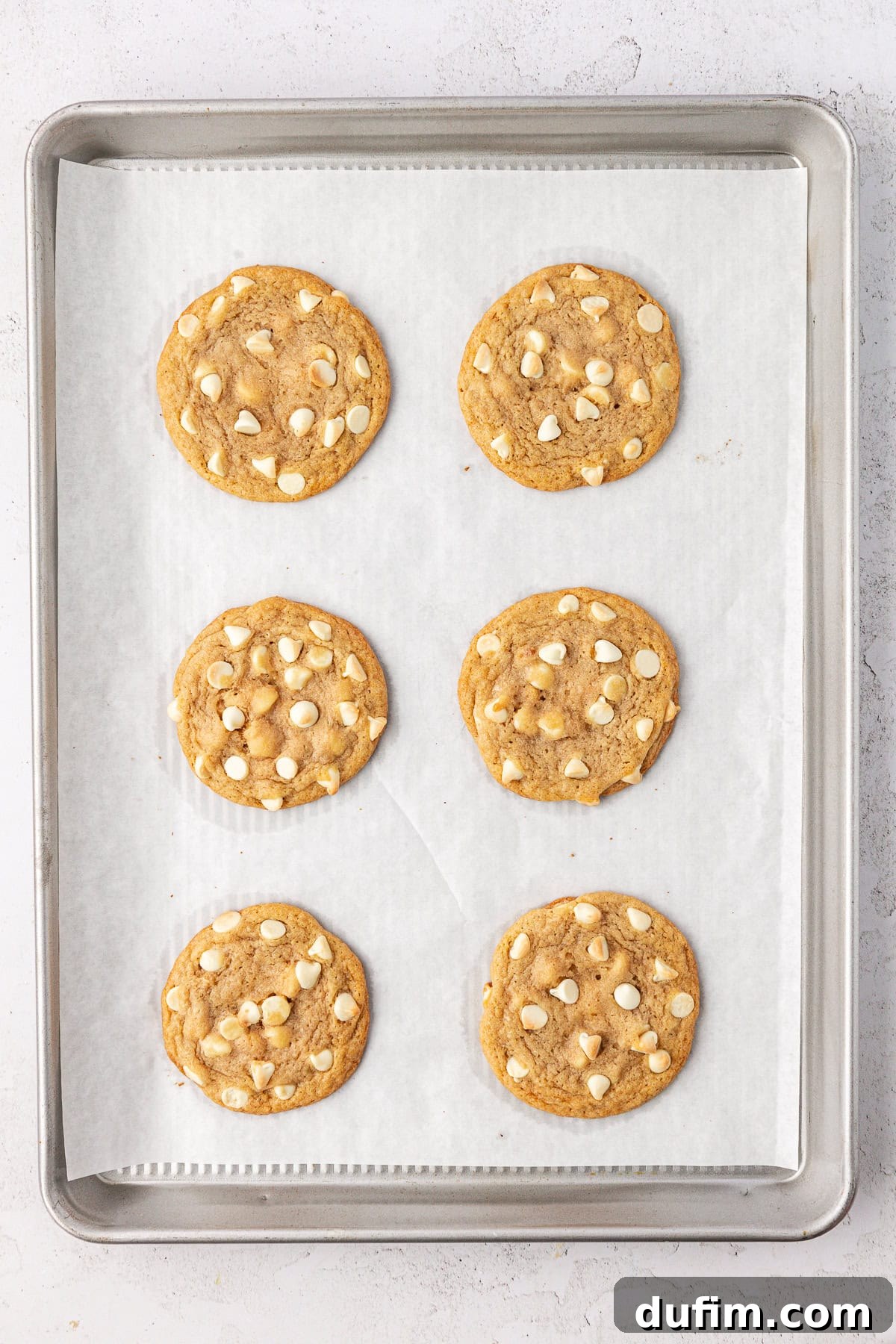 Overhead view of two rows of three white chocolate chips cookies on a baking sheet lined with white parchment paper