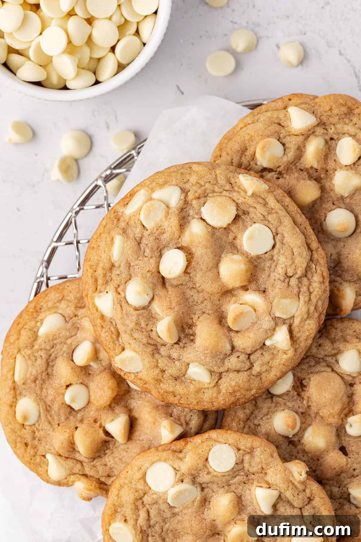 Overhead view of white chocolate chip cookies piled on a wire rack with white chocolate chips in a small white bowl beside it and scattered around